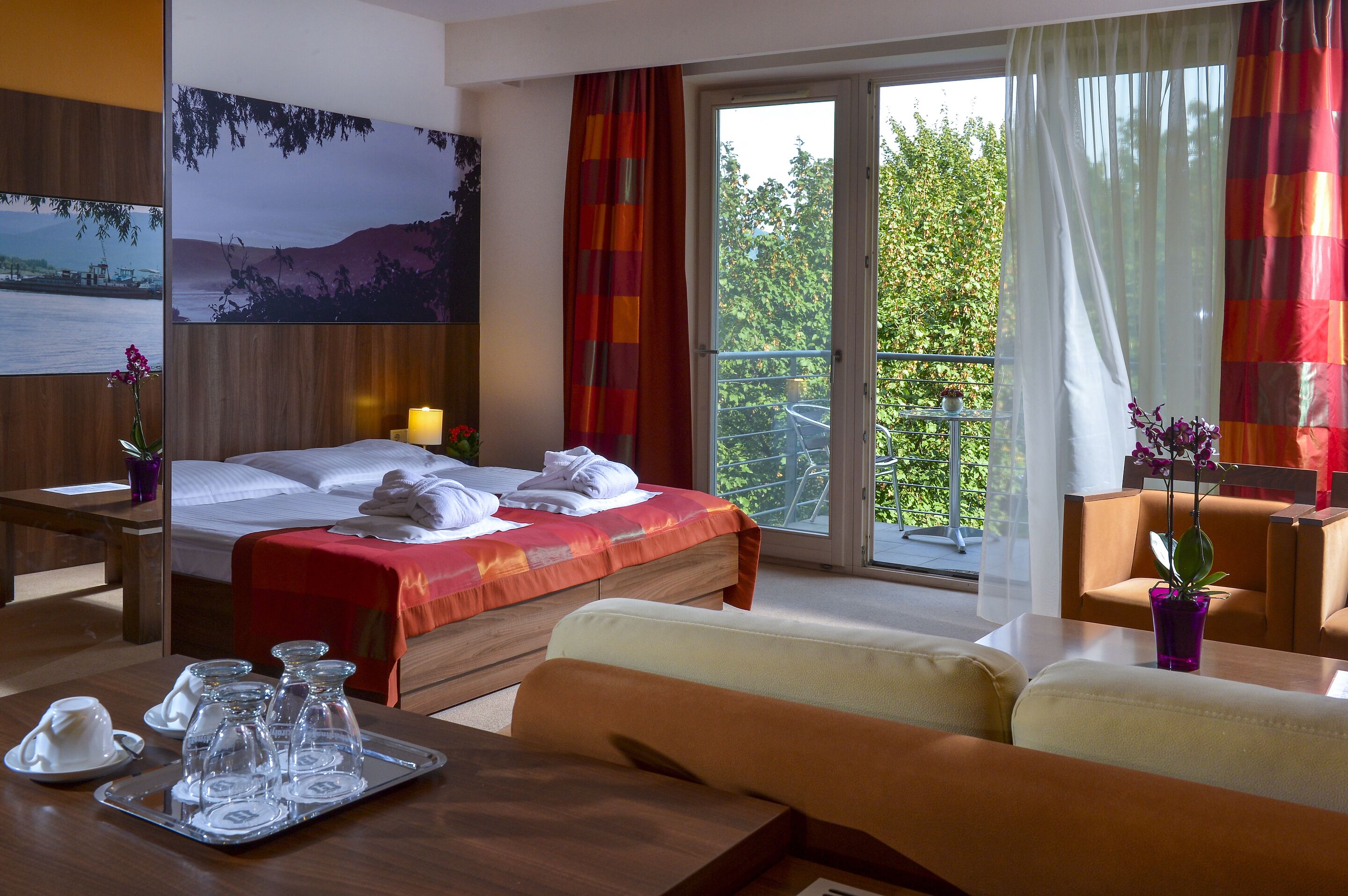 A hotel room with a bed, a sofa, a dining table, and a balcony with a view of trees.