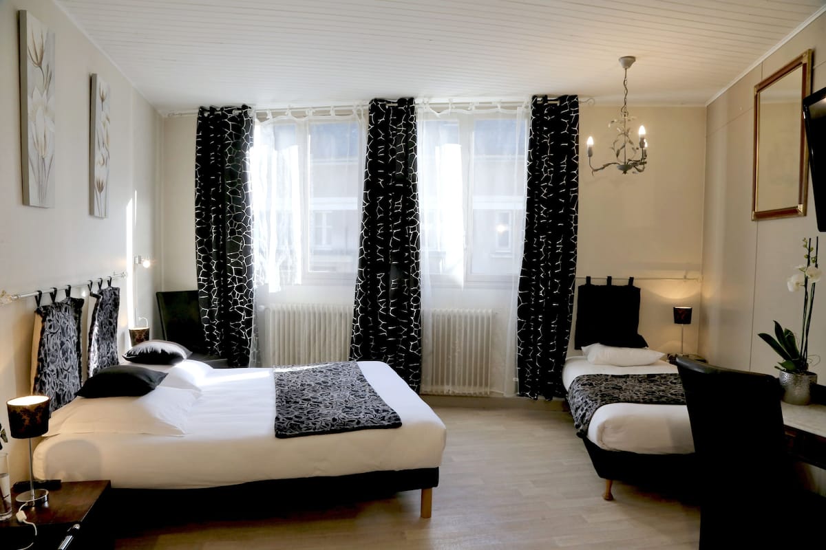 Ein Hotelzimmer mit einem großen Bett, einem kleineren Bett, einem Schreibtisch, einem Stuhl, einer Kristallleuchter und einer Topfpflanze.