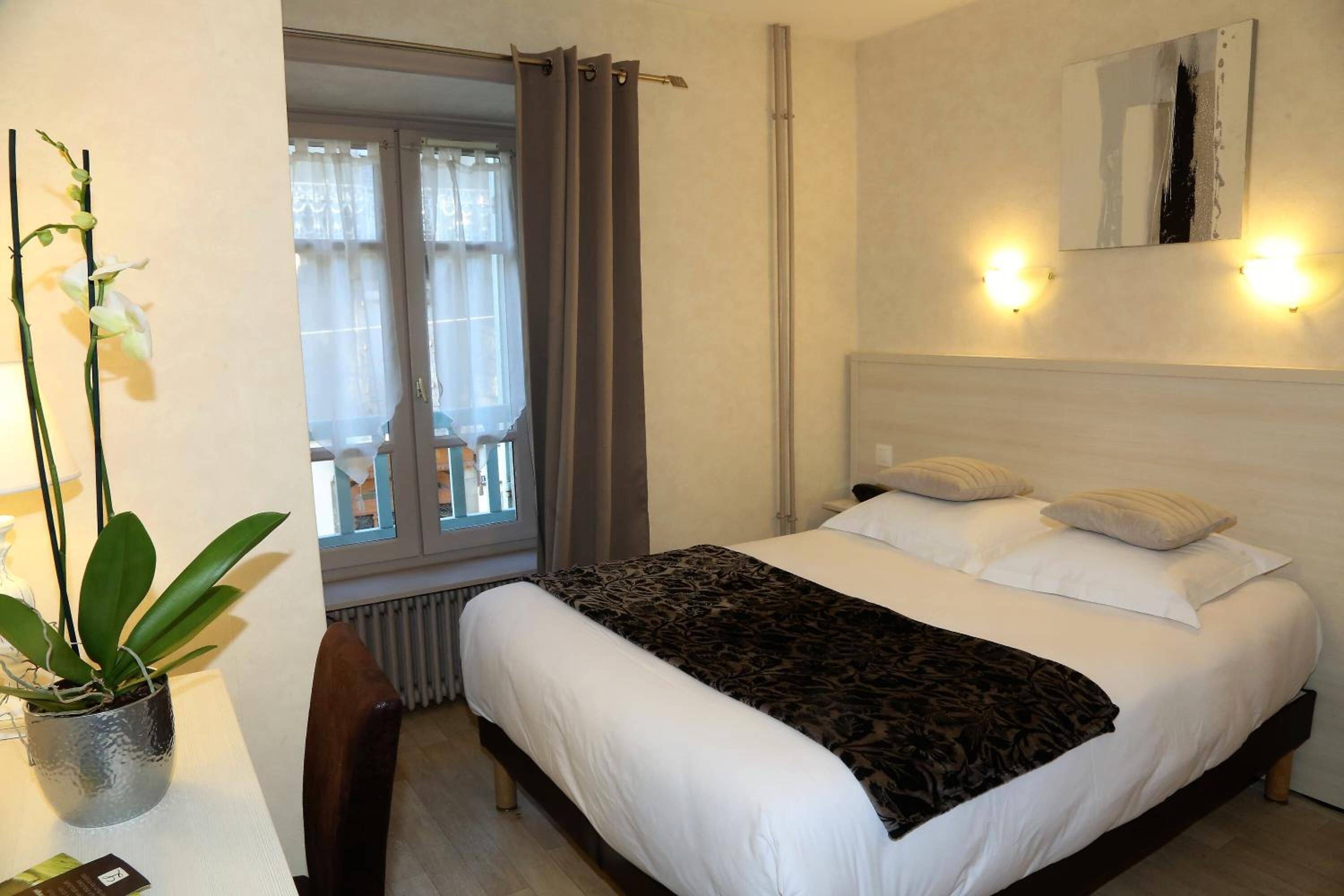 Quarto de hotel com cama, planta em vaso, cadeira, janela com cortinas e luminária na parede.