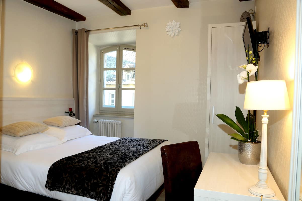 Ein Hotelzimmer mit einem Bett, einem Schreibtisch mit Lampe, einer Zimmerpflanze und einem Fenster mit Vorhängen.