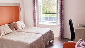 Uma cama de casal com cabeceira acolchoada, duas mesas de cabeceira, uma escrivaninha com cadeira e uma janela com cortinas.