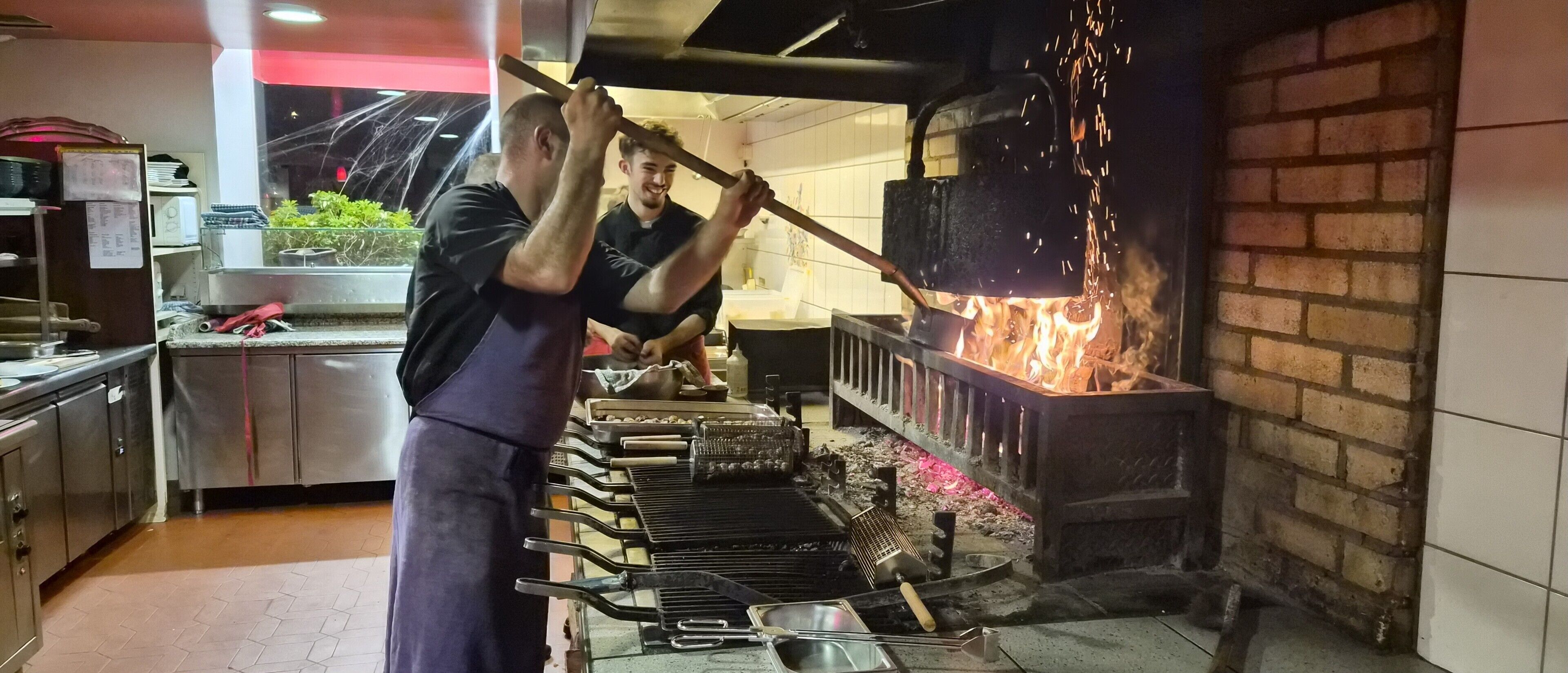 Une cuisine équipée d’un four en briques, d’appareils électroménagers en acier inoxydable, et où une personne utilise un outil à long manche.
