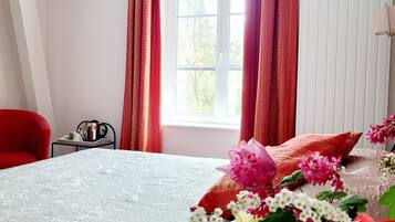 Un dormitorio con una cama, una silla roja, una mesita con objetos y una ventana con cortinas.