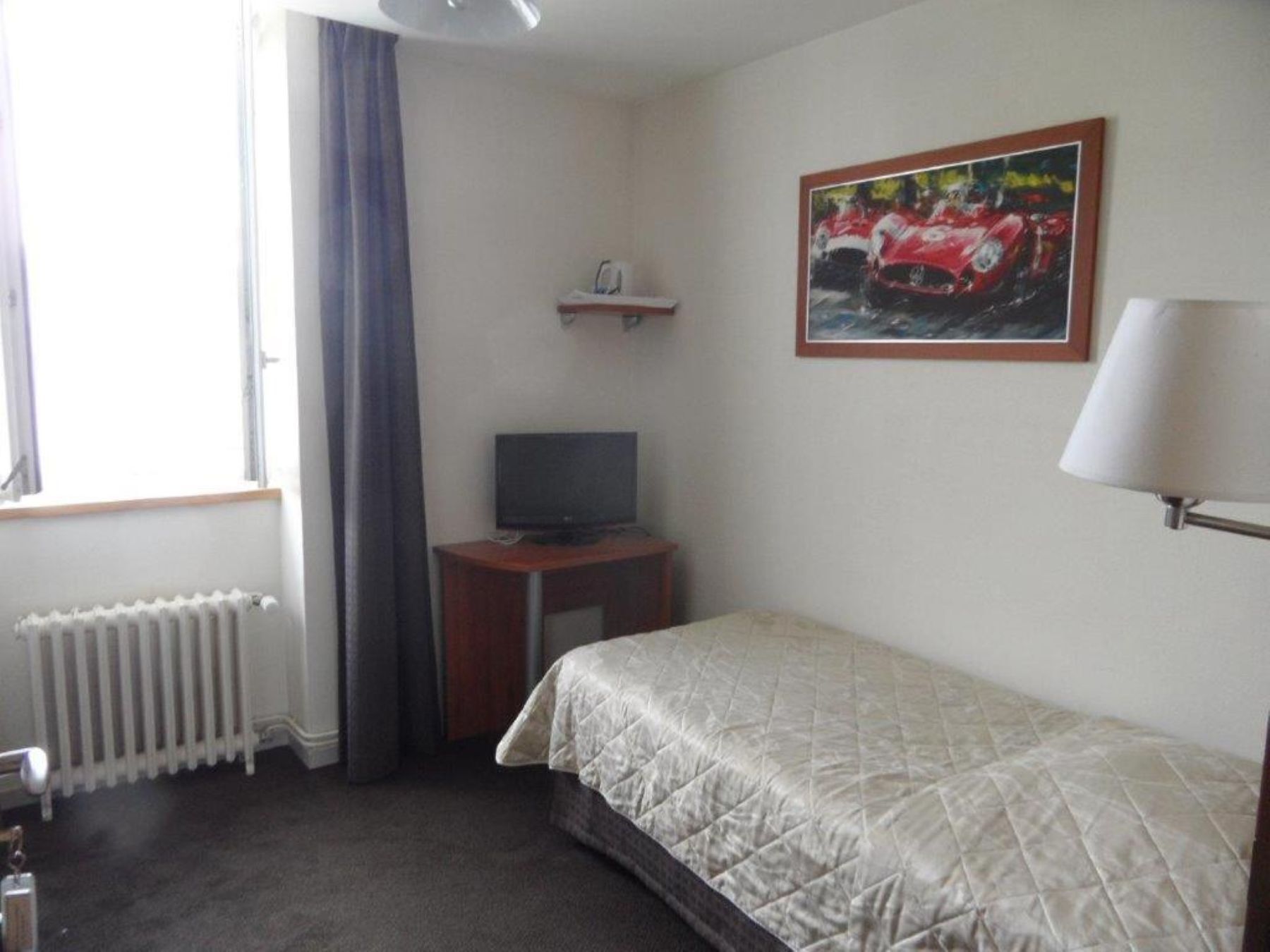 A hotel room with a bed, a television on a wooden stand, a radiator, and a window with curtains.