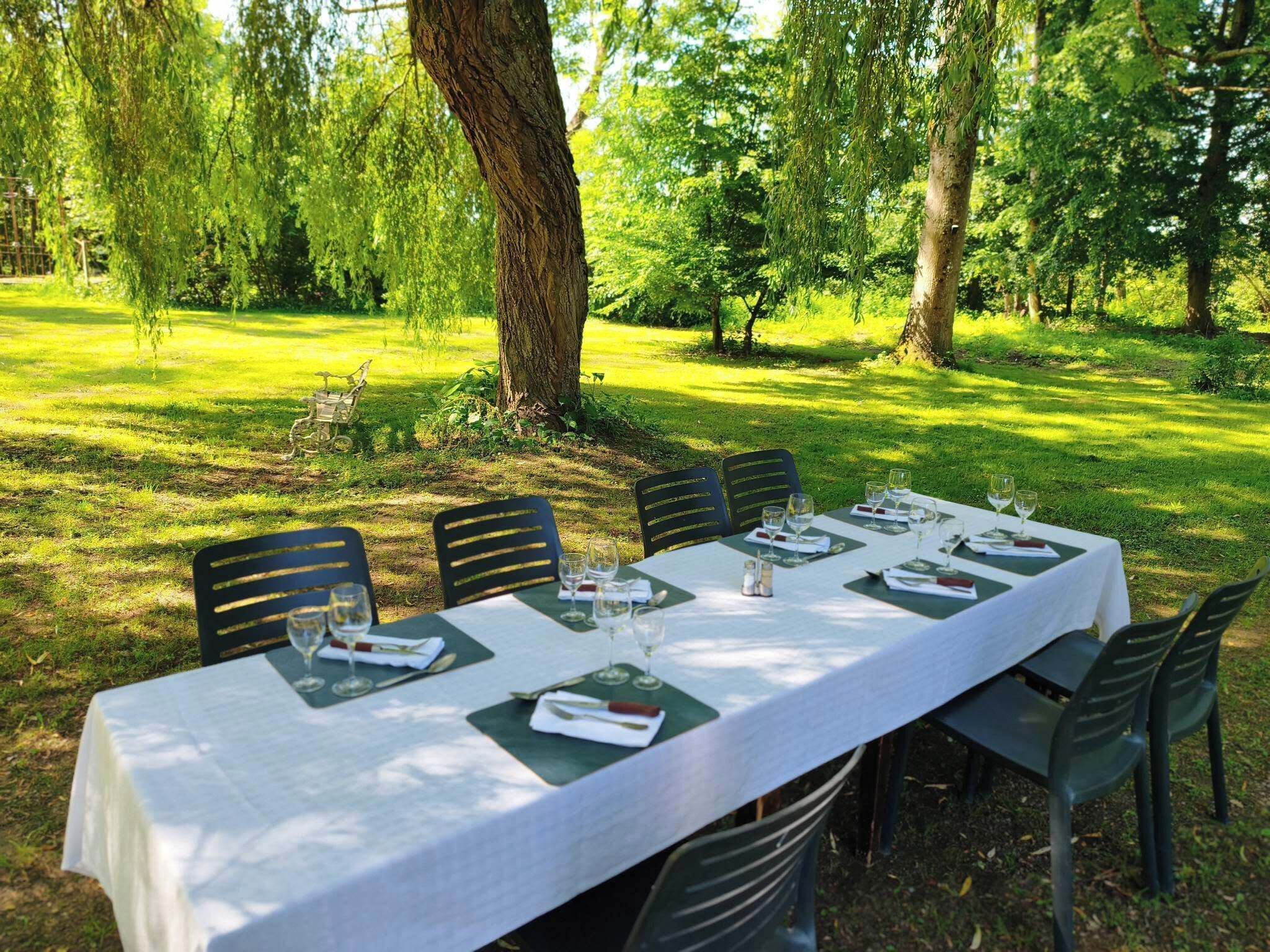 An outdoor dining area with a table set for six, surrounded by chairs, in a lush green park setting.