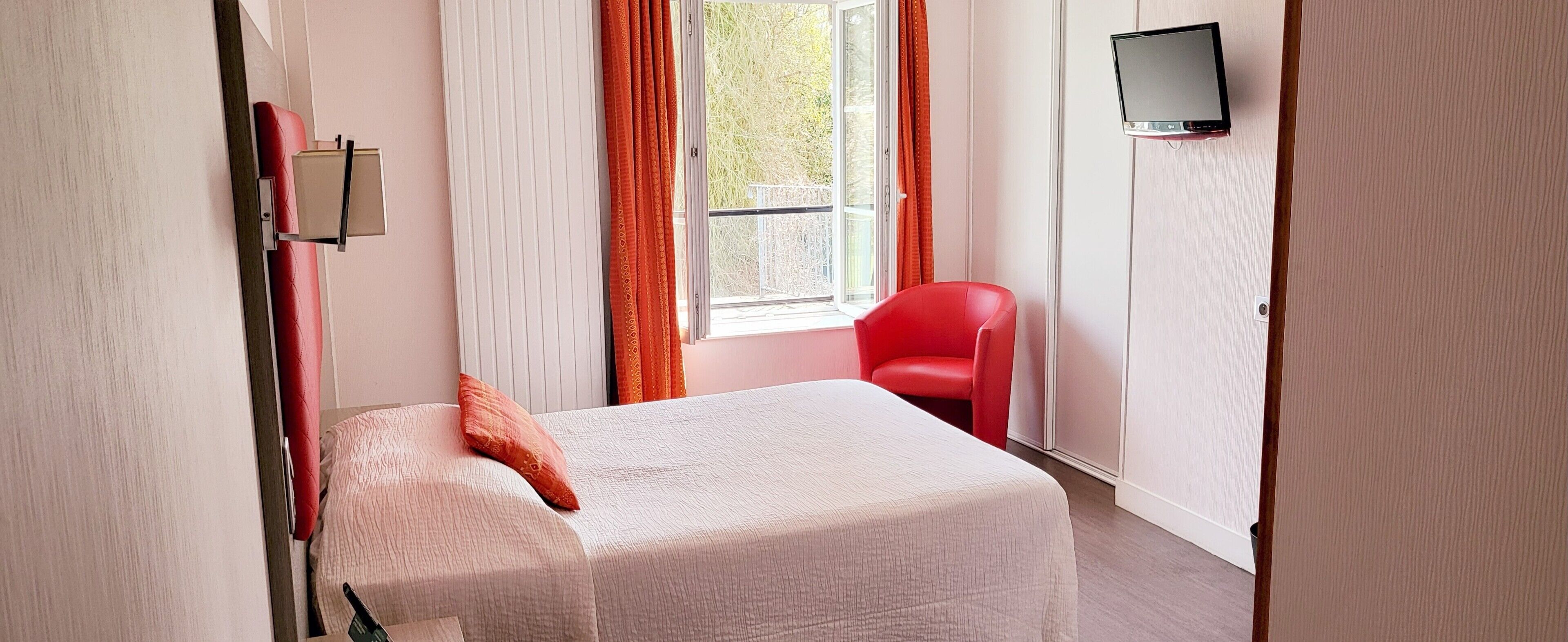 A bedroom with a bed, a red chair, a TV mounted on the wall, and a window with curtains.