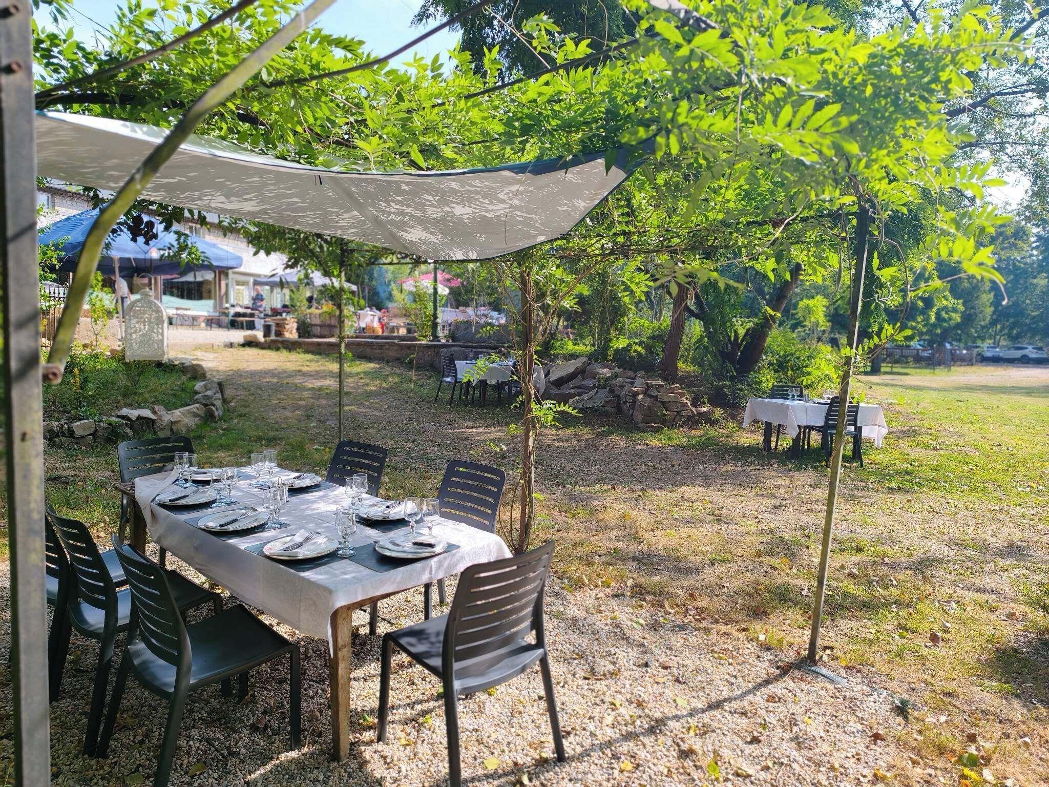 An outdoor dining area with tables set for two, surrounded by trees and a stone wall.