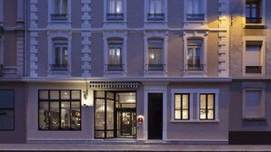 En bygning med flere etager i klassisk arkitekturstil, med en central indgang med glasdøre, flere vinduer med balkoner, og en lygtepæl der oplyser facaden.