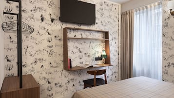 A hotel room with a bed, a desk with a mirror, a TV mounted on the wall, and a window with curtains.