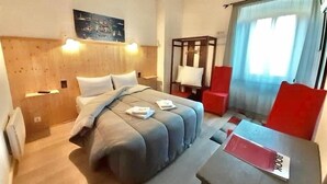A hotel room with a wooden headboard, a bed with a grey duvet, a wooden nightstand, a red chair, a table with a red folder, and a window with curtains. - Hotel Bayle (Belcaire)