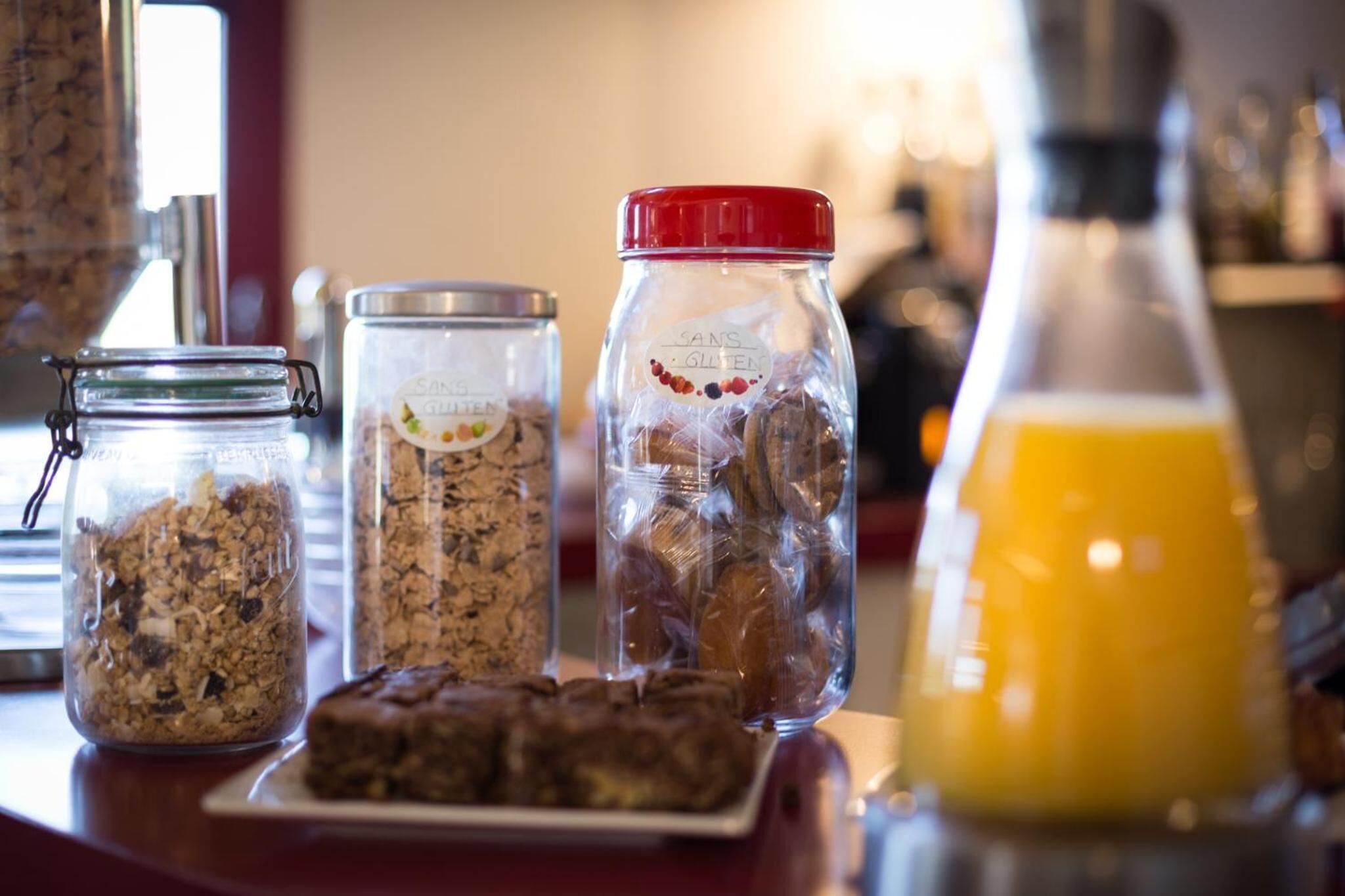 Tres frascos de vidrio con etiquetas, un plato con brownies y una botella de jugo de naranja sobre una encimera.