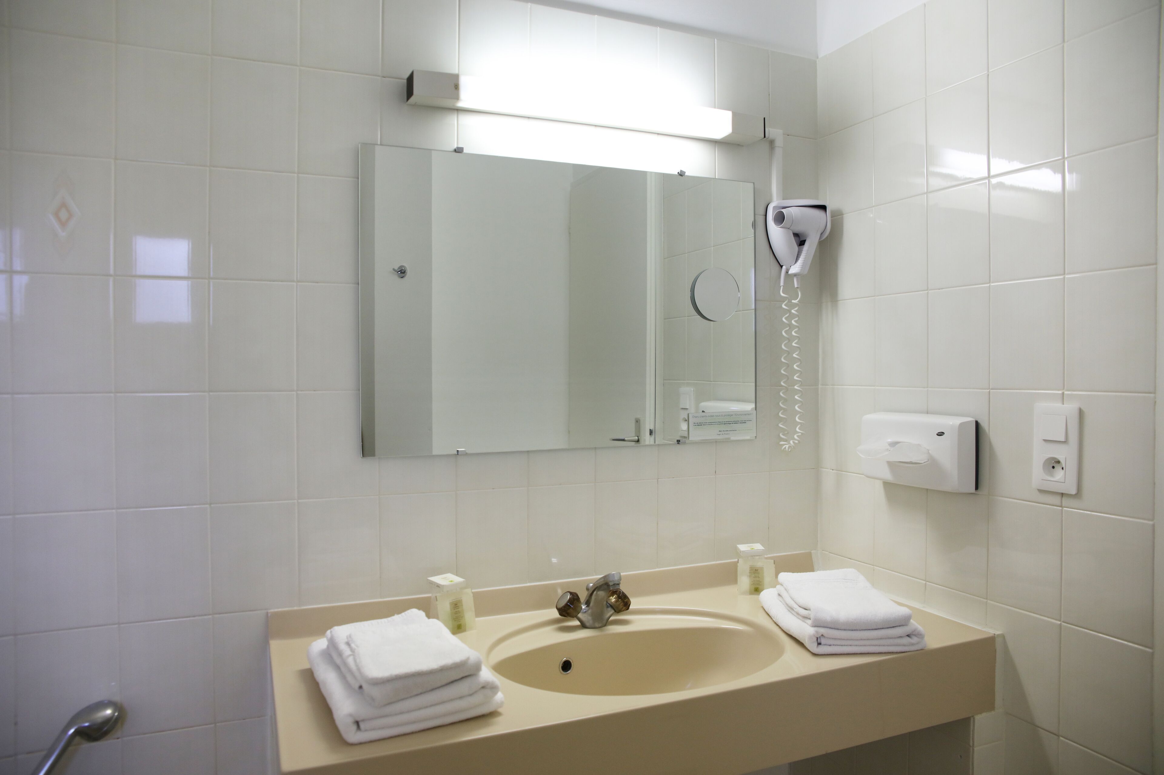 Ein Badezimmer mit Waschbecken, Spiegel, Handtuchspender und einem an der Wand montierten Fön.