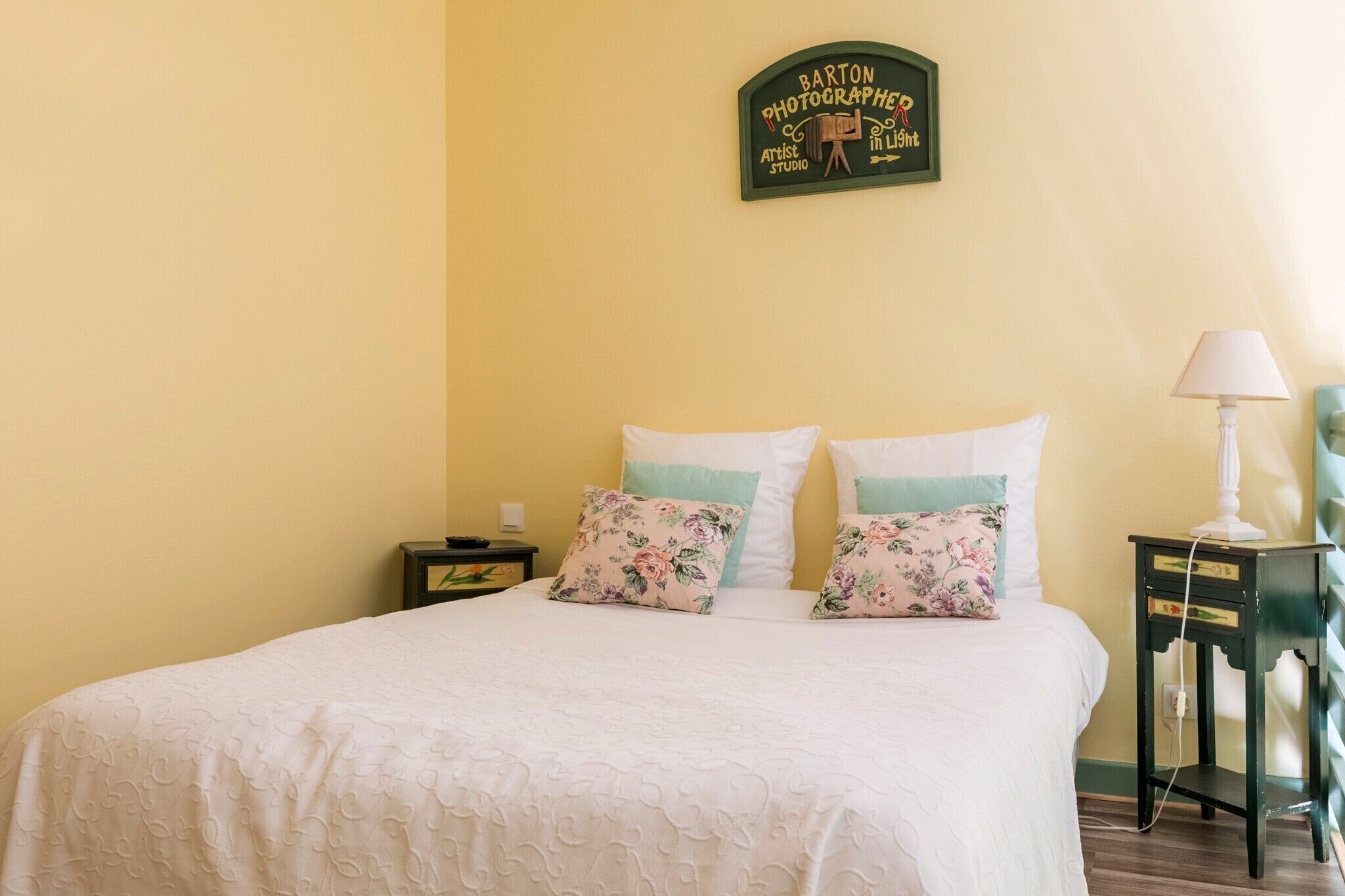 A neatly made bed with floral pillows, a small bedside table with a lamp, and a wall sign above the bed.
