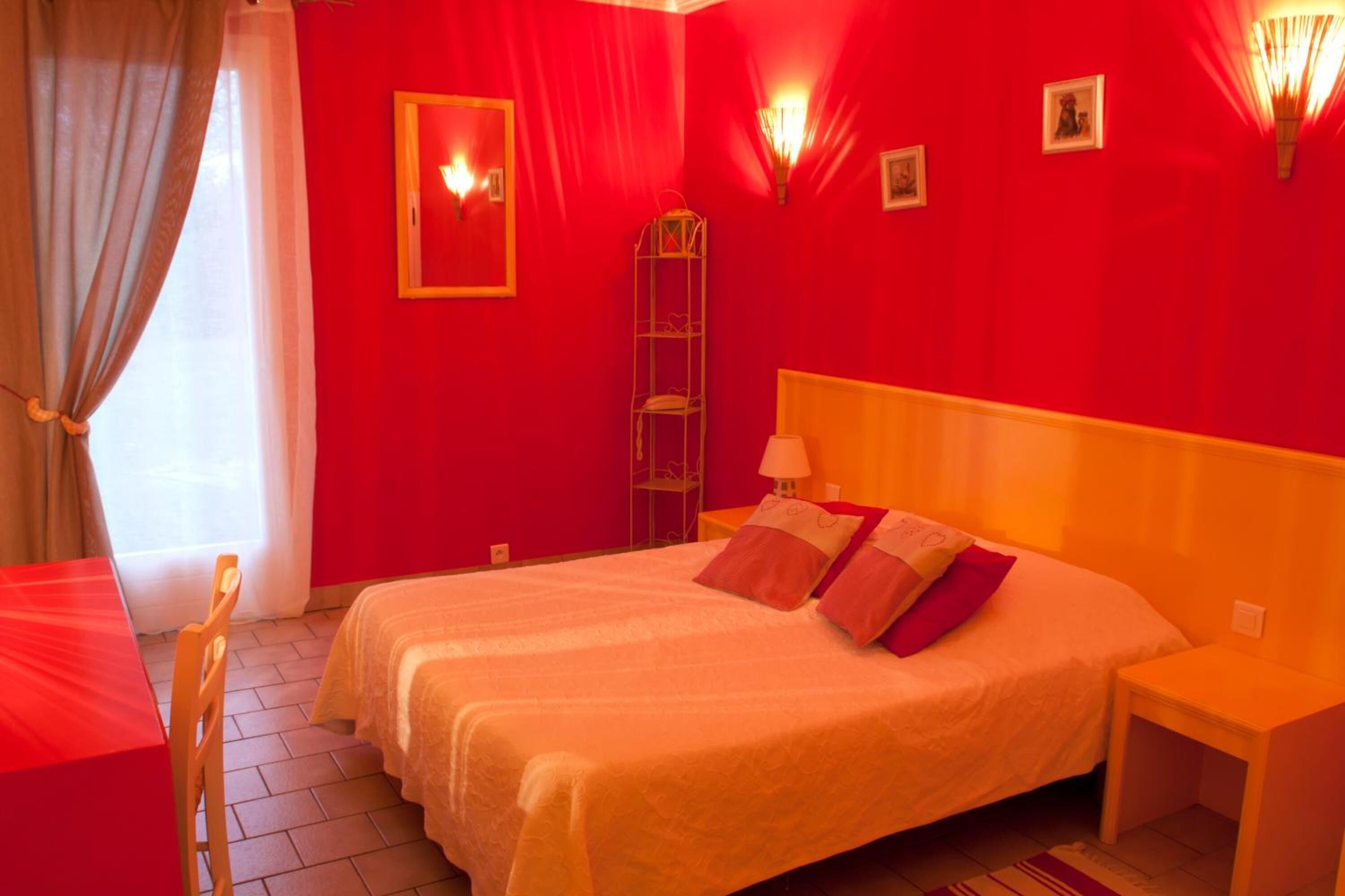 A hotel room with a red wall, a bed with white and pink bedding, a wooden chair, a bedside table, and a floor lamp.