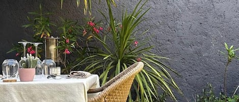 An outdoor dining area with a table set for two, surrounded by wicker chairs, potted plants, and a textured wall in the background.