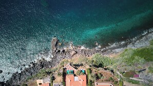 Vista aerea di una proprietà costiera con diversi edifici, un litorale roccioso e acque blu cristalline.