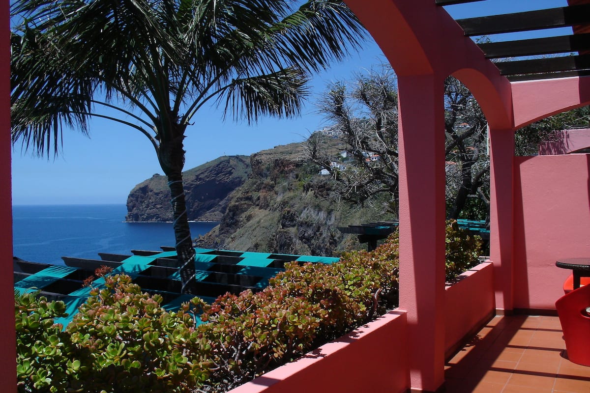 A terrace with a view of the sea, a palm tree, and a mountain in the background.