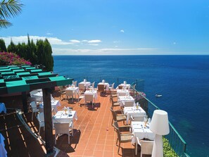 Une terrasse agrémentée de tables et de chaises, offrant une vue dégagée sur la mer et permettant d’apercevoir un bateau au loin.