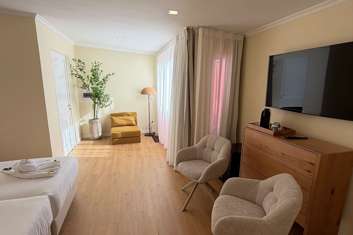 A hotel room with a bed, two armchairs, a wooden dresser, a television, a plant, and a window with curtains.