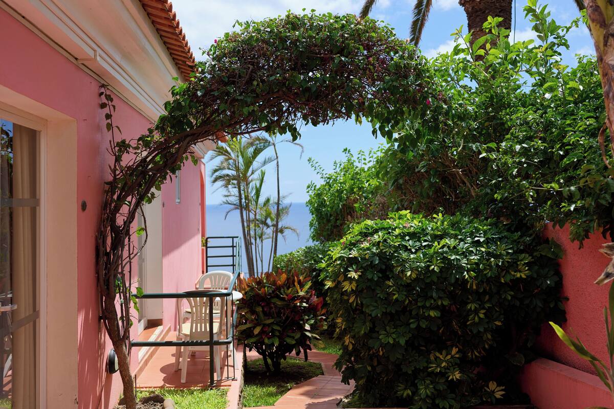 A pink building with a covered pathway, a white chair, and lush greenery.