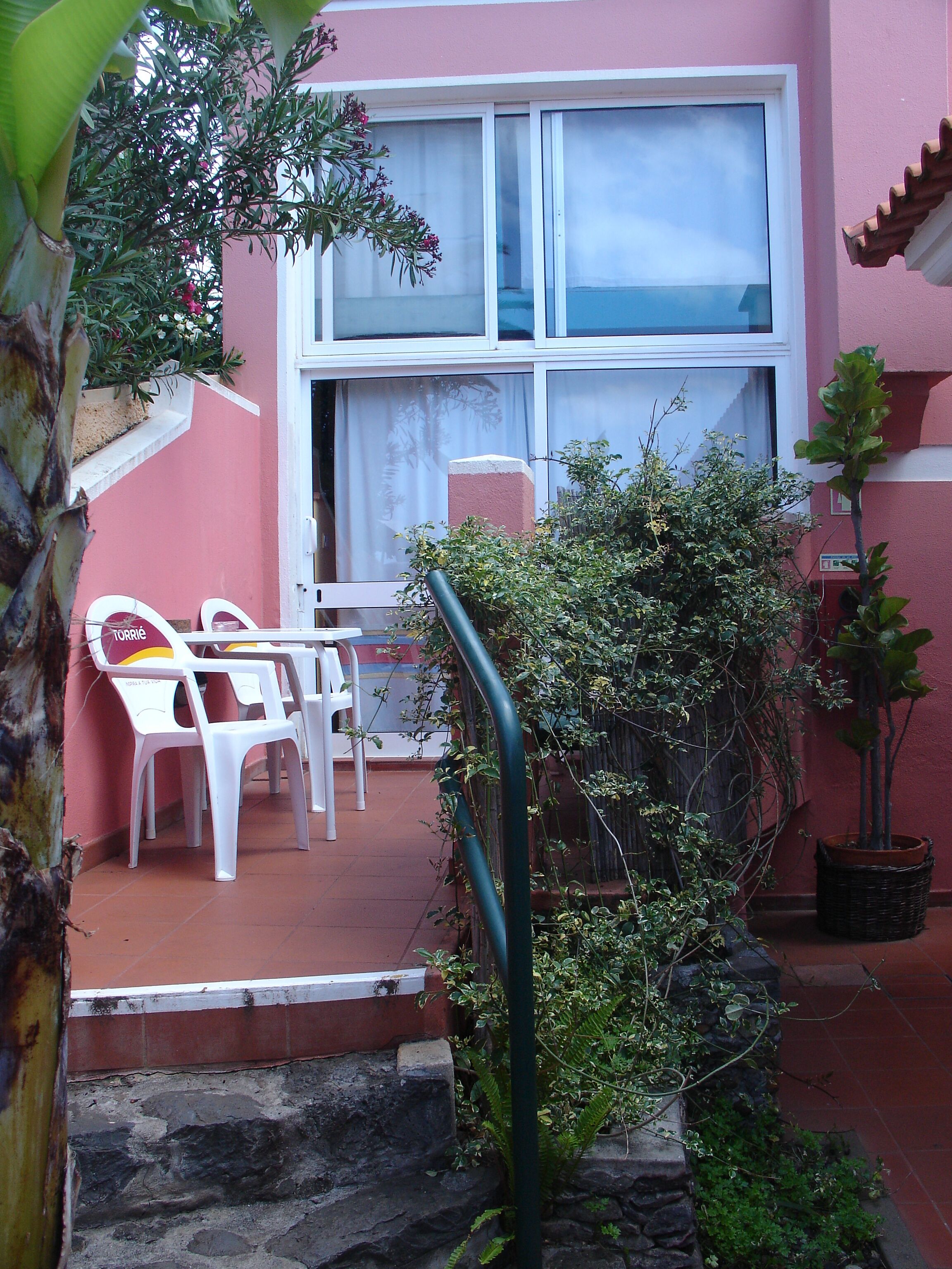 A small patio area with white plastic chairs and a table, a potted plant, and a staircase leading to a building with a pink exterior.