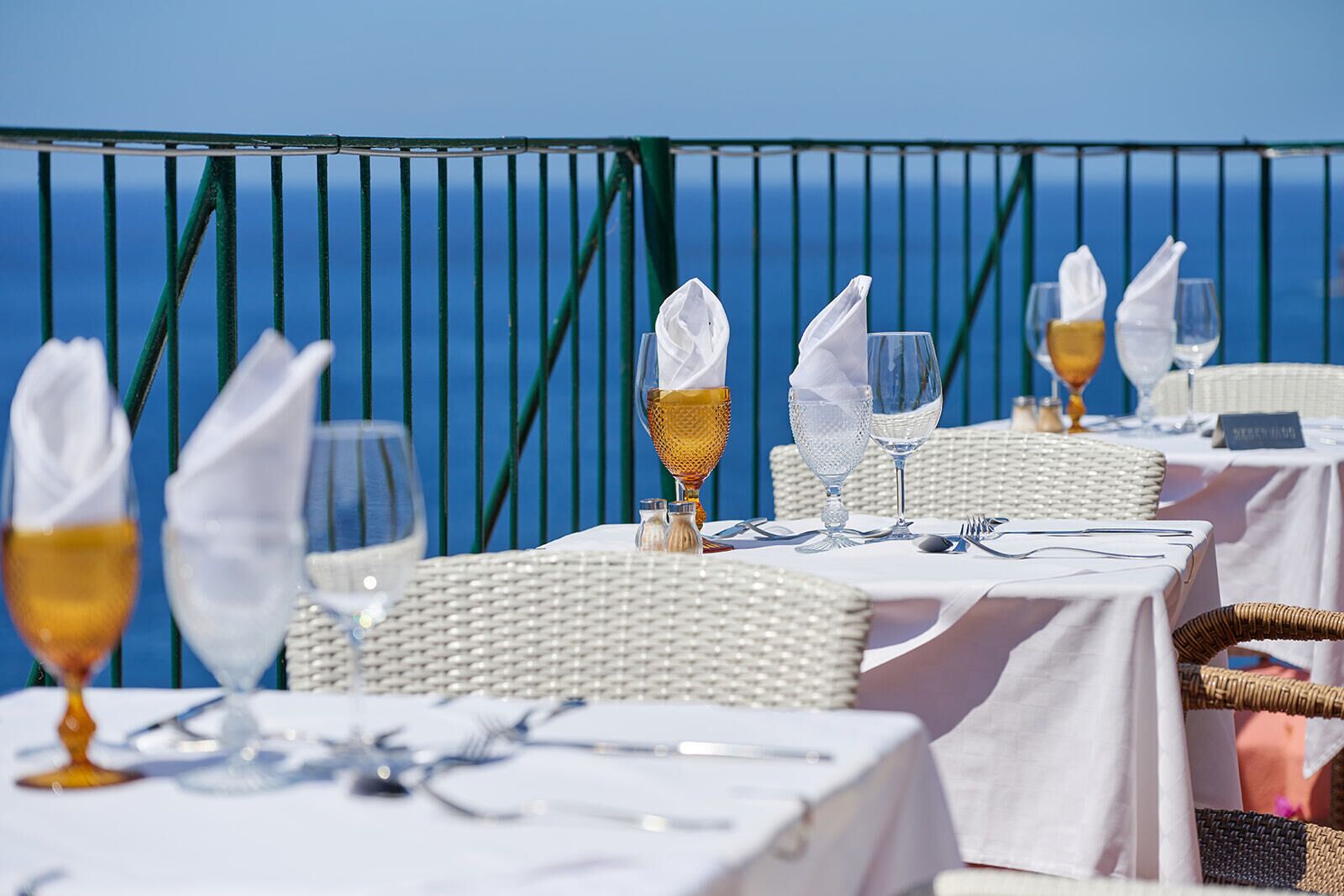 Mesa lista para comer con manteles blancos y sillas de mimbre, con vista al océano azul.