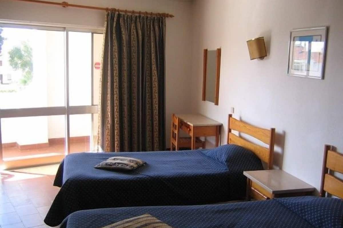 Ein Zimmer mit zwei Betten, einem Schreibtisch, einem Stuhl und einem fensterbepflanzten Fenster.