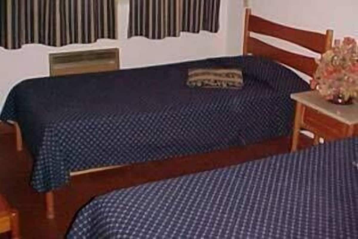 Ein Schlafzimmer mit zwei Einzelbetten, einem hölzernen Kopfteil, einem Nachttisch mit Lampe und einem Fenster mit Vorhängen.