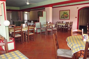 A dining area with multiple tables and chairs, a ceiling fan, and a kitchen in the background.