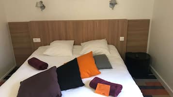 Habitación de hotel con cabecera de madera, dos lámparas fijadas a la pared, cama con ropa blanca y un cesto de basura negro.