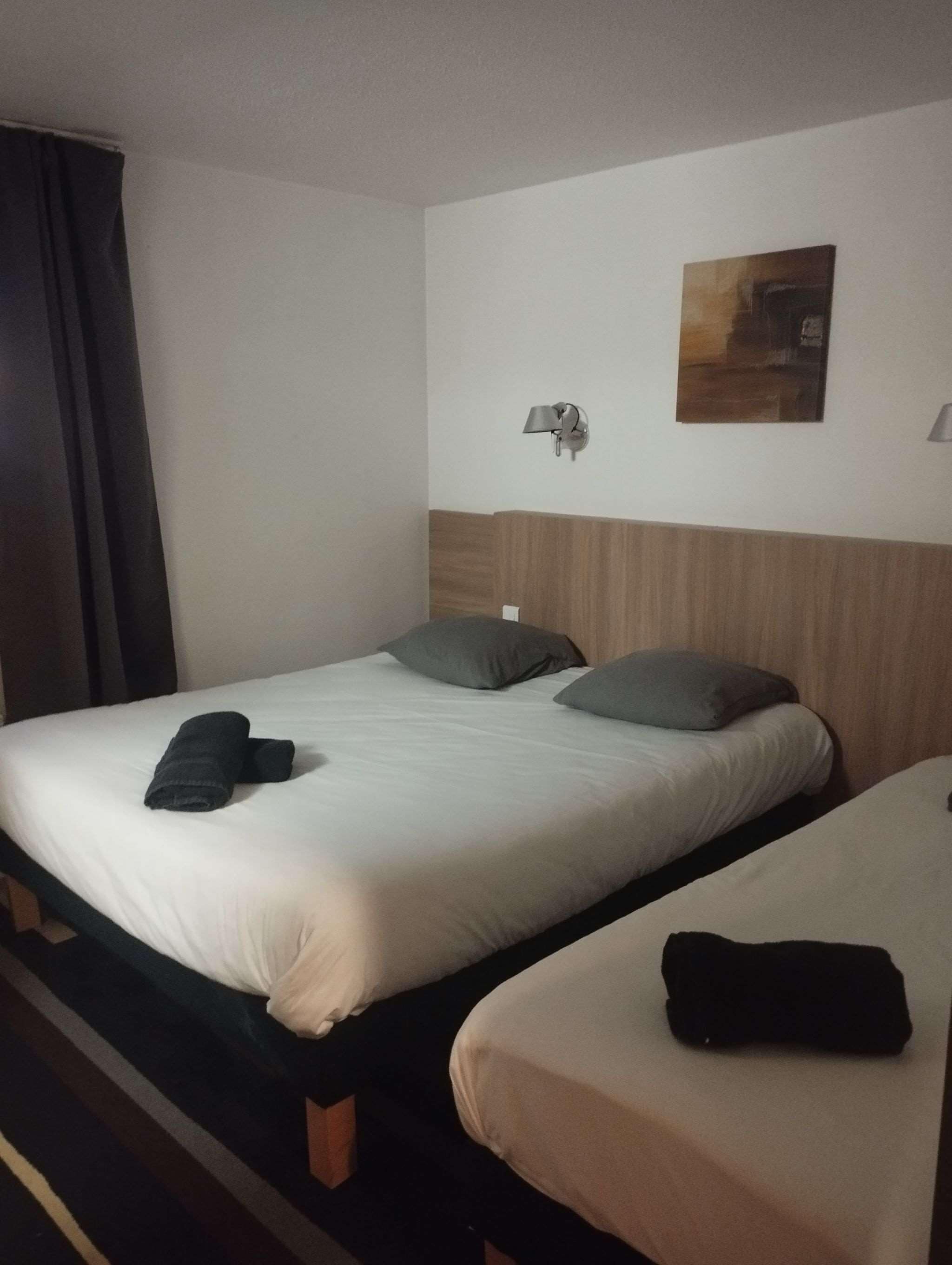 Habitación de hotel con dos camas individuales, cada una con una almohada y una toalla enrollada. Hay una lámpara de pared y un cuadro sobre las camas.