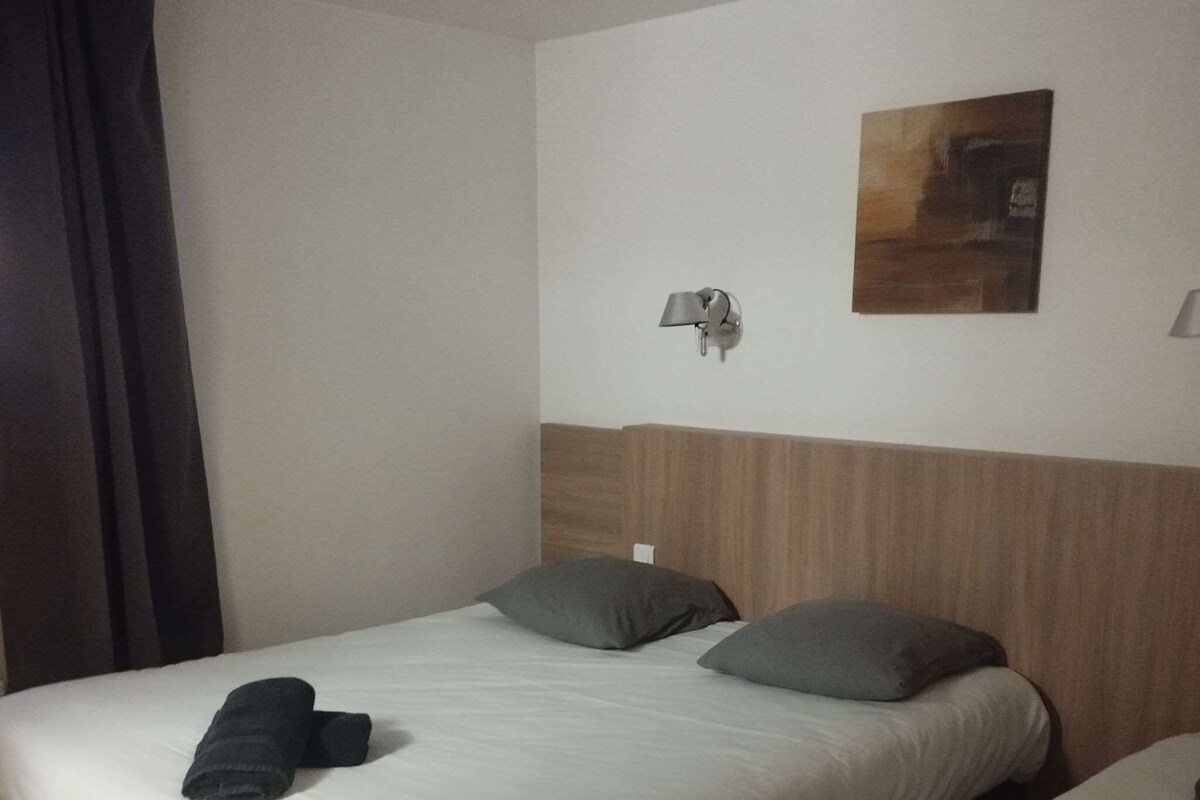 A hotel room with two single beds, each with a pillow and a rolled towel. There is a wall-mounted lamp and a painting above the beds.