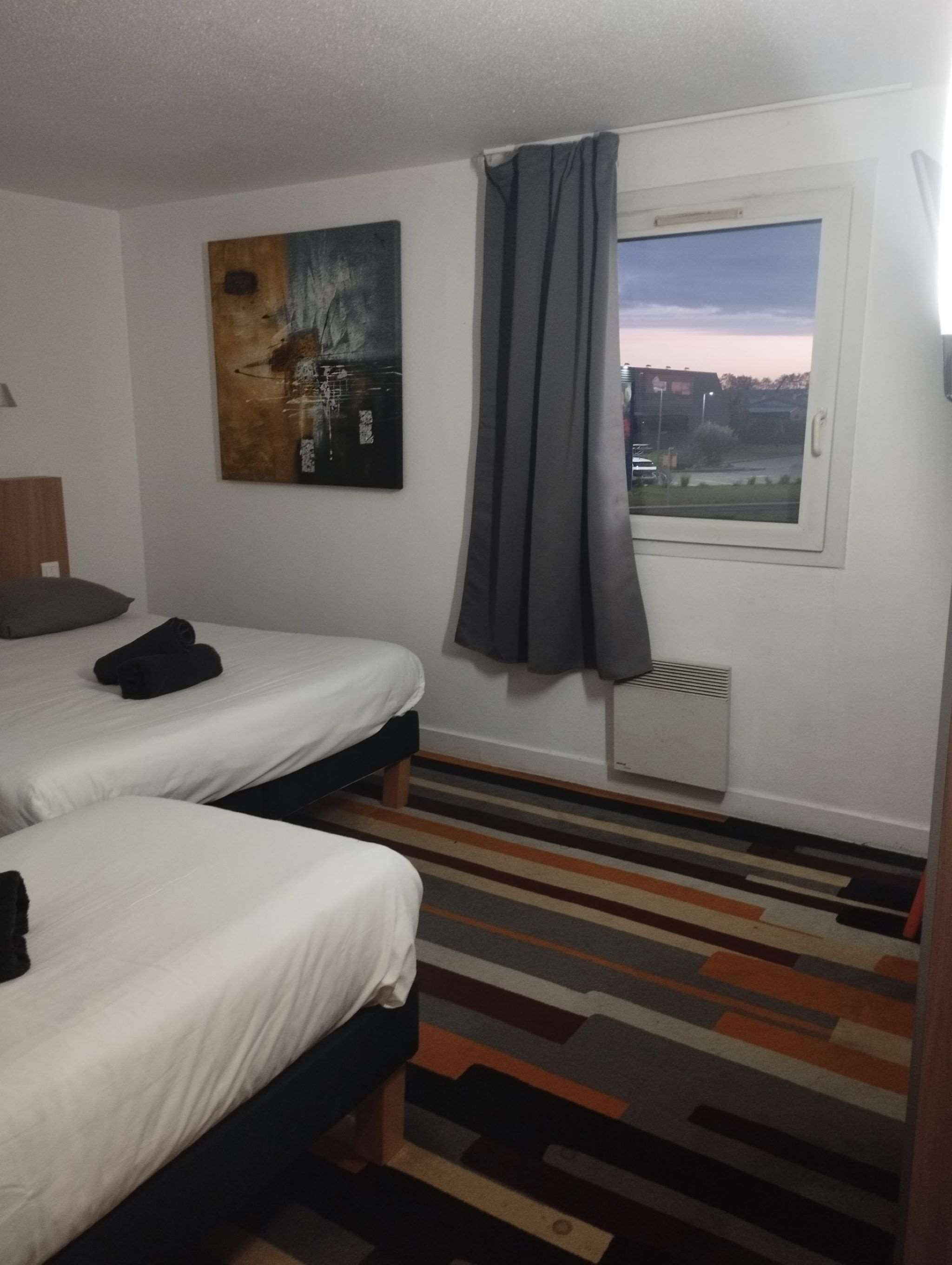 Une chambre d’hôtel avec deux lits, une fenêtre avec des rideaux, un radiateur et une moquette rayée aux couleurs vives.