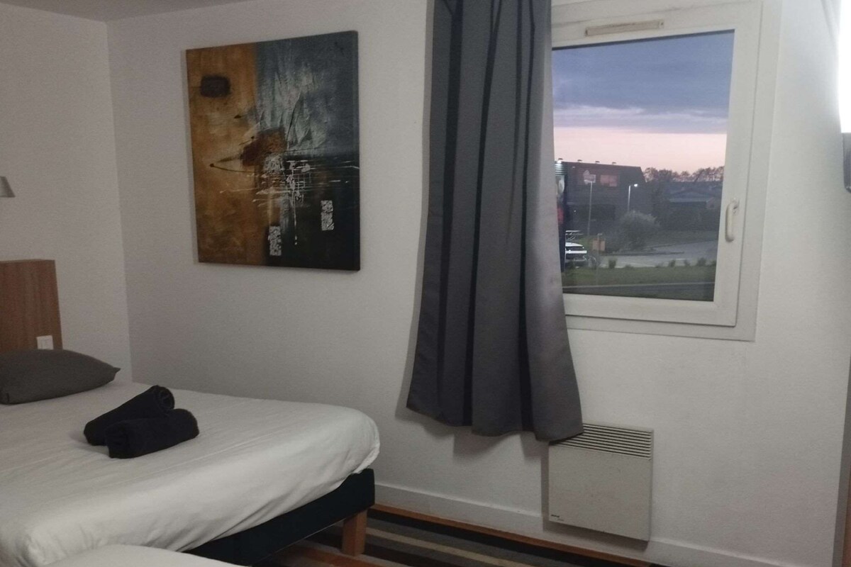 A hotel room with two beds, a window with curtains, a radiator, and a colorful striped carpet.