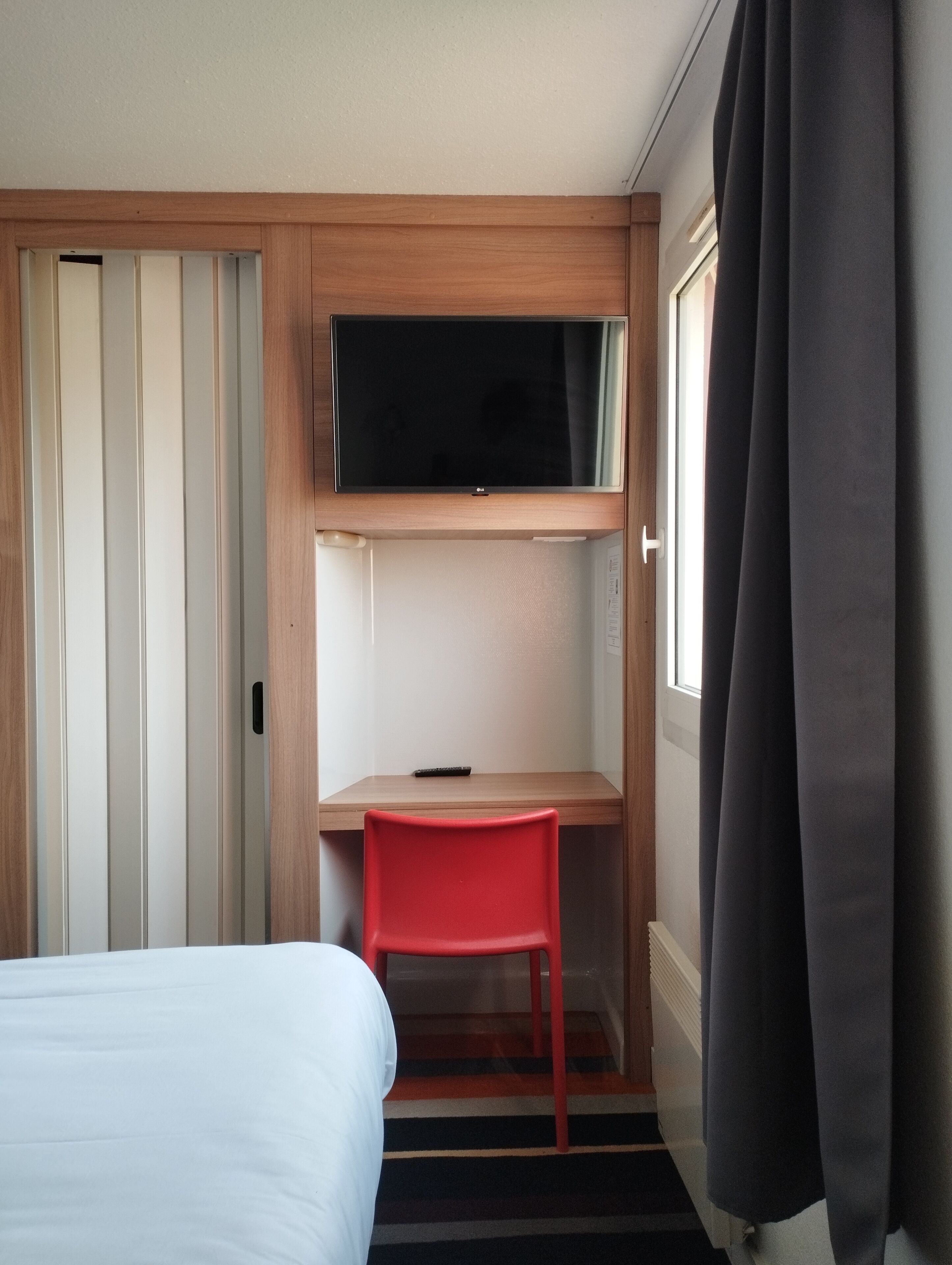 A hotel room with a bed, a desk with a chair, a TV mounted above the desk, and a window with curtains.