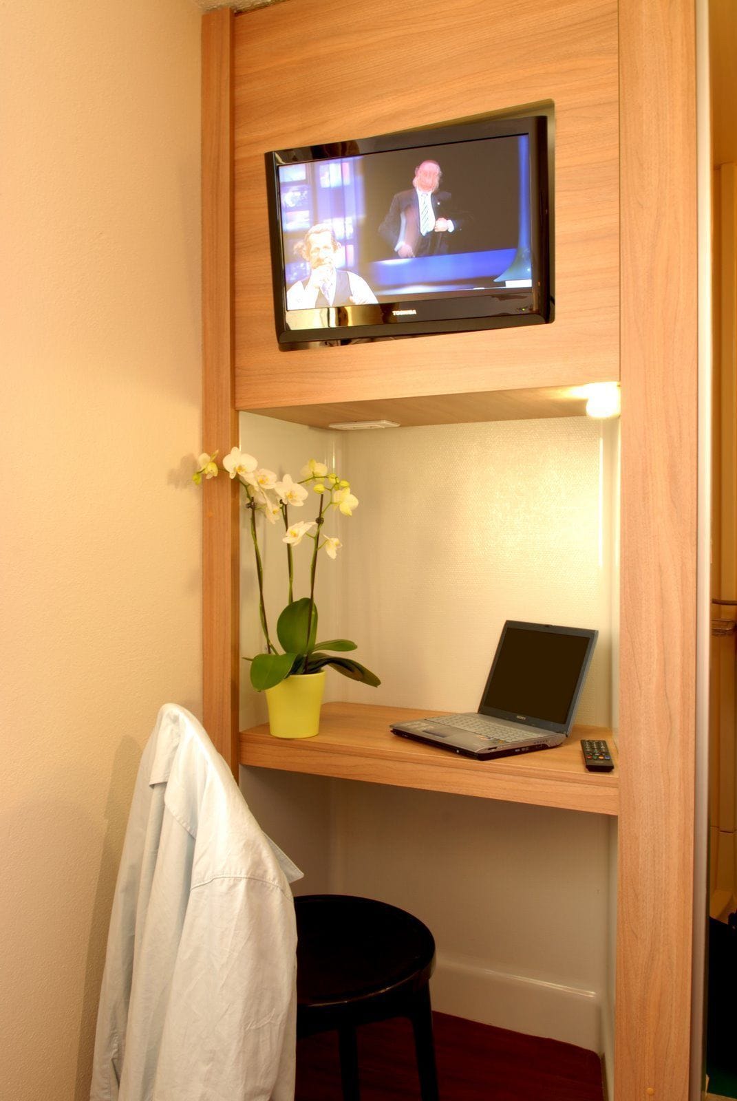 Una cama litera con un televisor montado sobre la cama superior, una computadora portátil en la inferior, una orquídea en maceta y una toalla blanca colgada al lado.