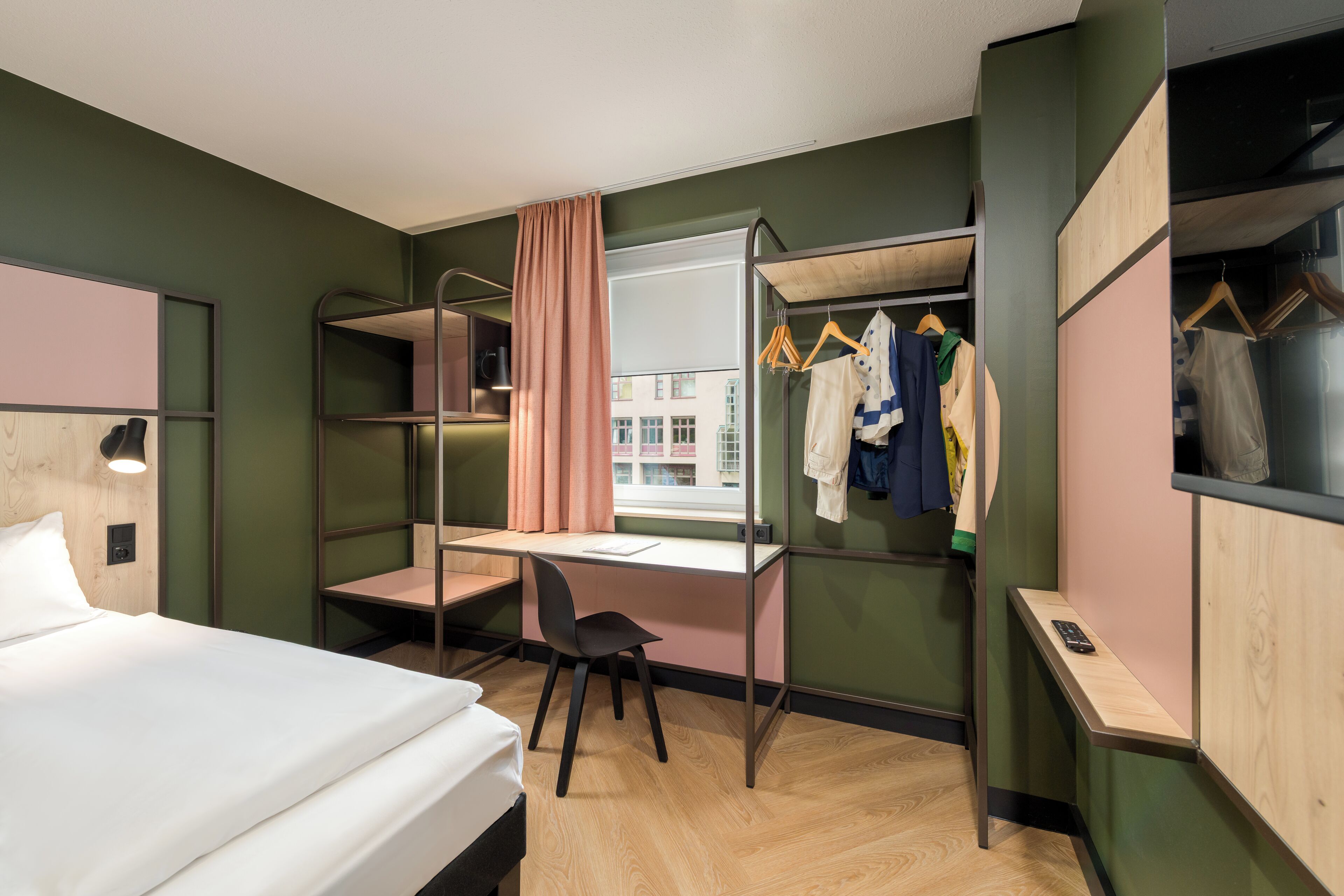 Habitación de hotel pequeña con literas, un escritorio, una silla y un armario con ropa colgada.