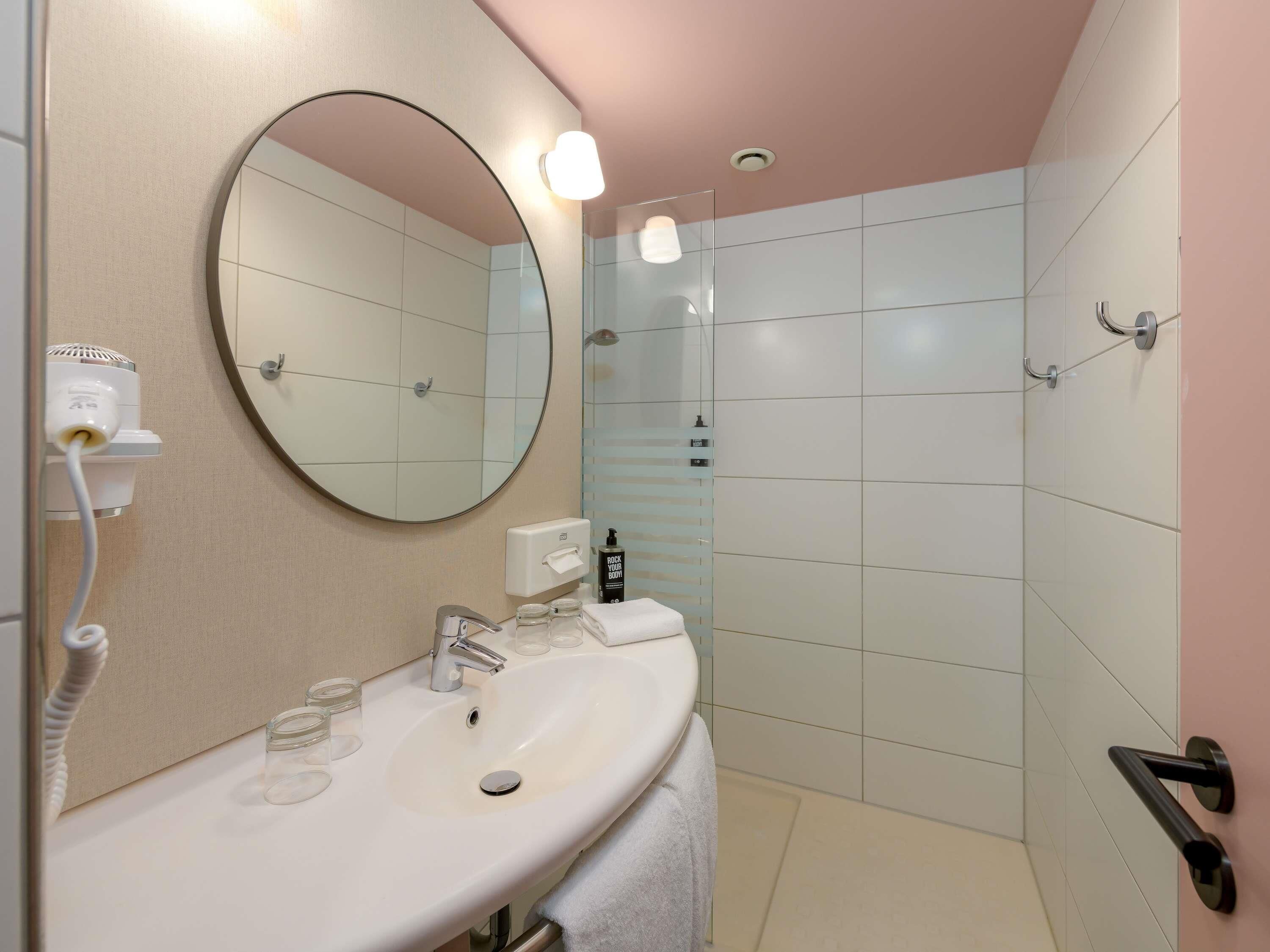Ein Badezimmer mit rundem Spiegel, Waschbecken, Handtuch, Duschkabine und Handtrockner.