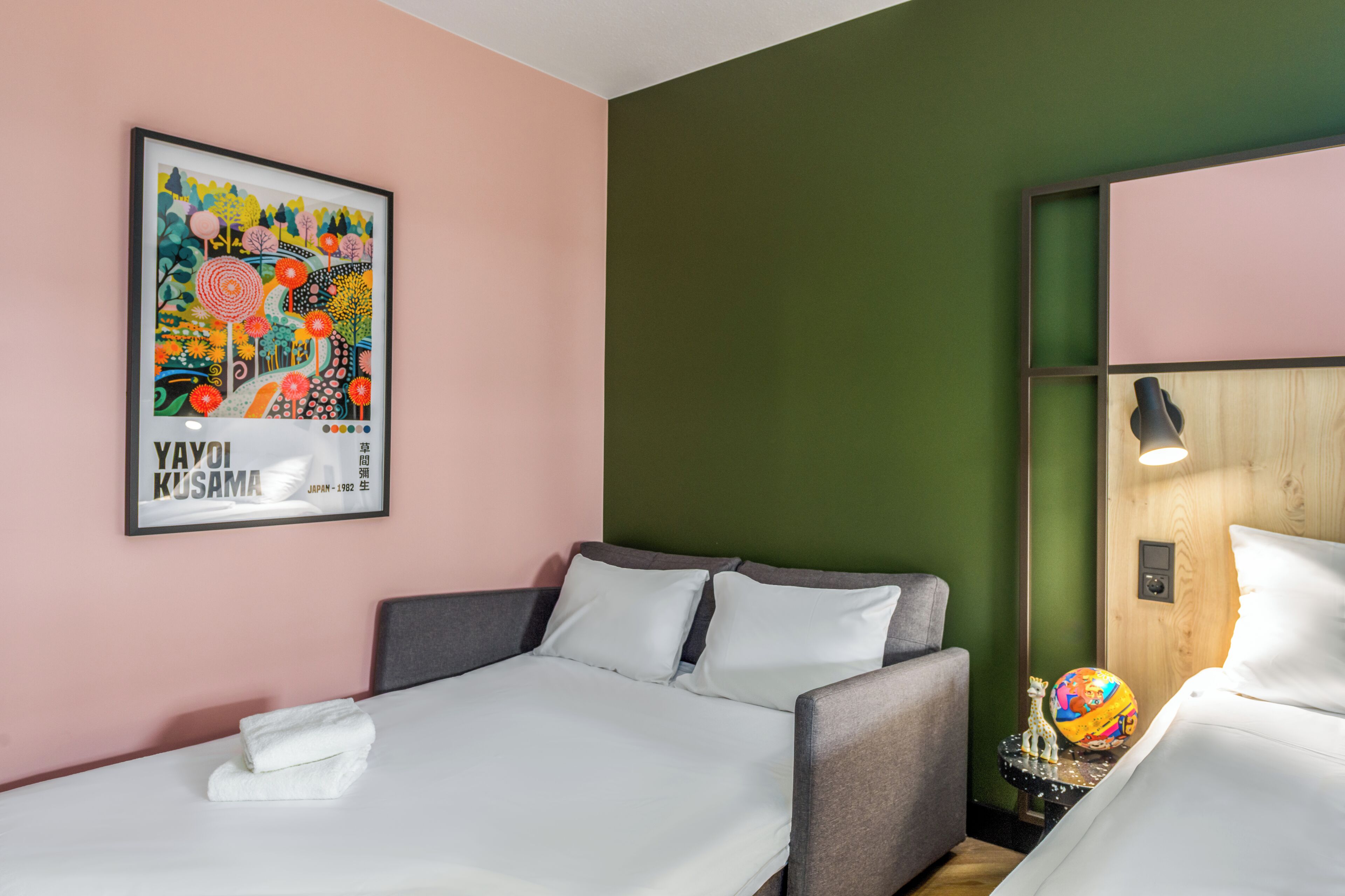 A hotel room with a bed, a sofa, a wall with a colorful poster, and a small table with a globe and a giraffe figurine.