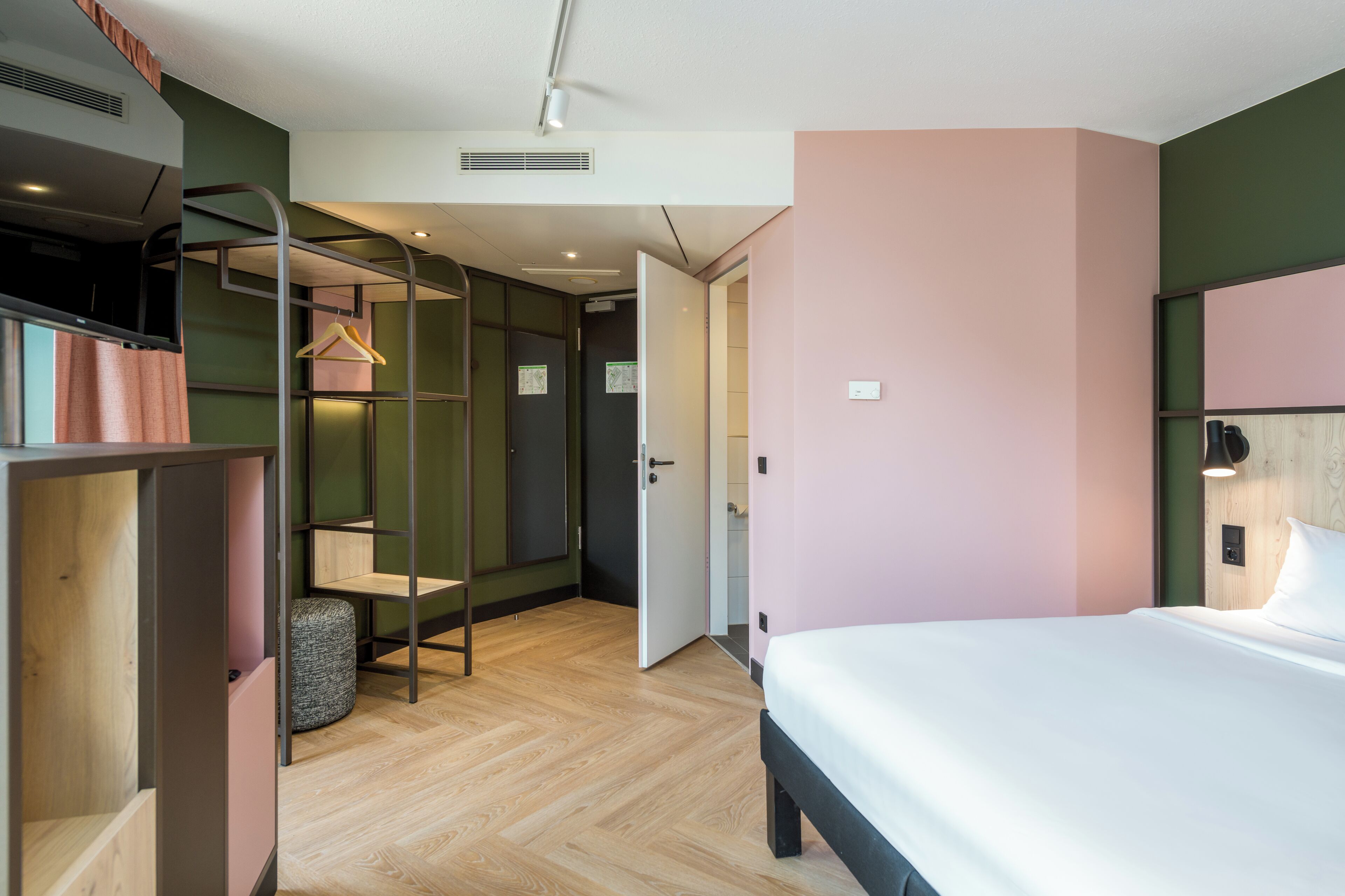 A modern hotel room with a bed, a wardrobe, a seating area with a chair, and a door leading to a bathroom.