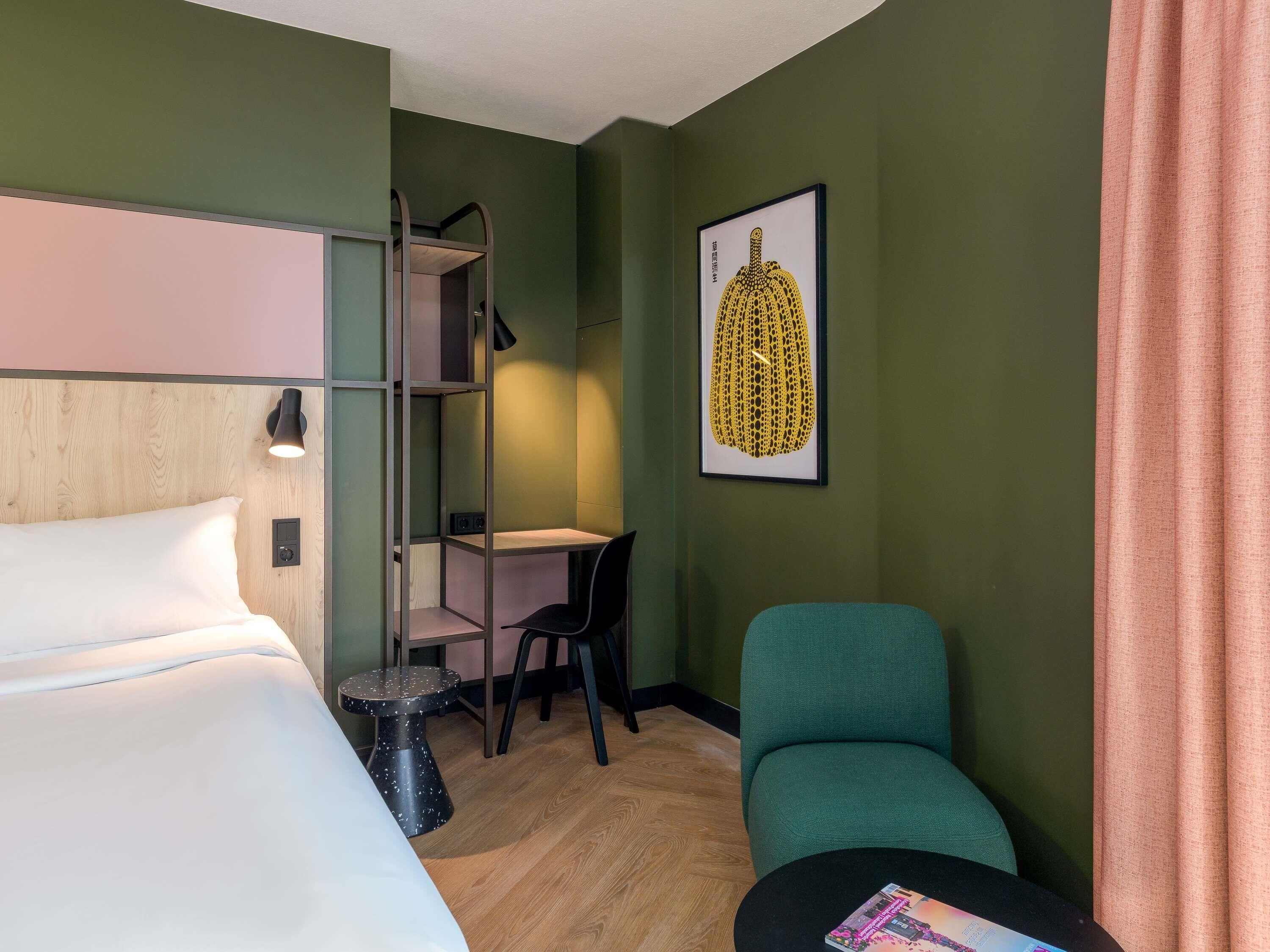 Une chambre d’hôtel équipée d’un lit, d’un bureau, d’une chaise, d’une petite table et d’une étagère fixée au mur.