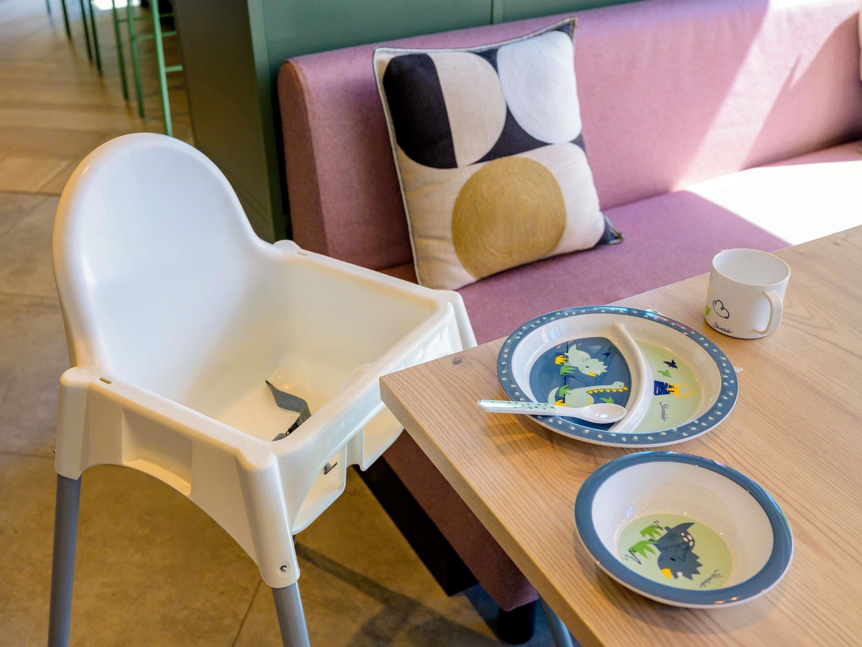 Ein Essbereich mit Hochstuhl, Tisch mit Tellern und Tasse sowie einer Couch mit gemusterten Kissen.