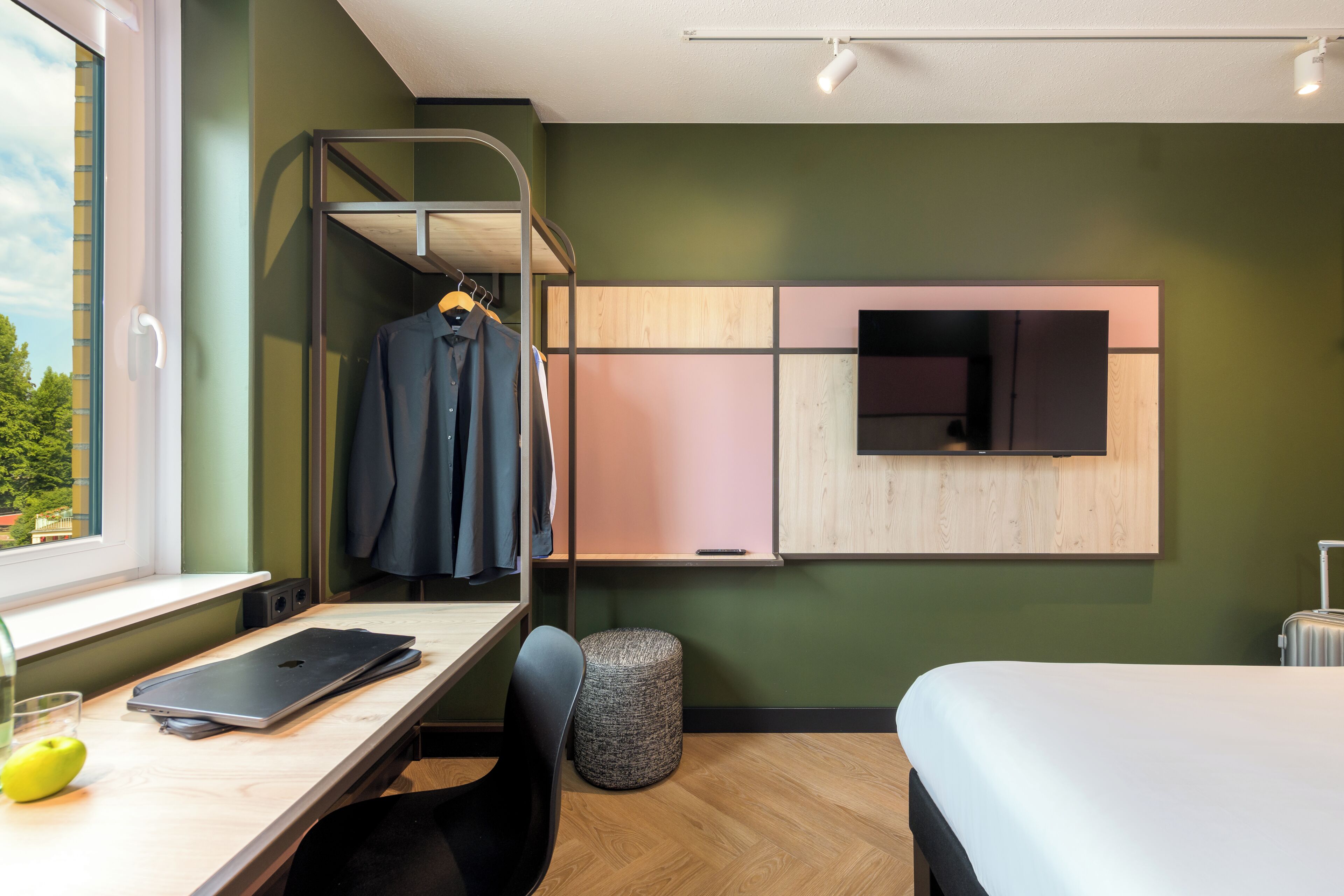 A hotel room with a bunk bed, a desk with a laptop, a chair, a small round ottoman, and a suitcase.