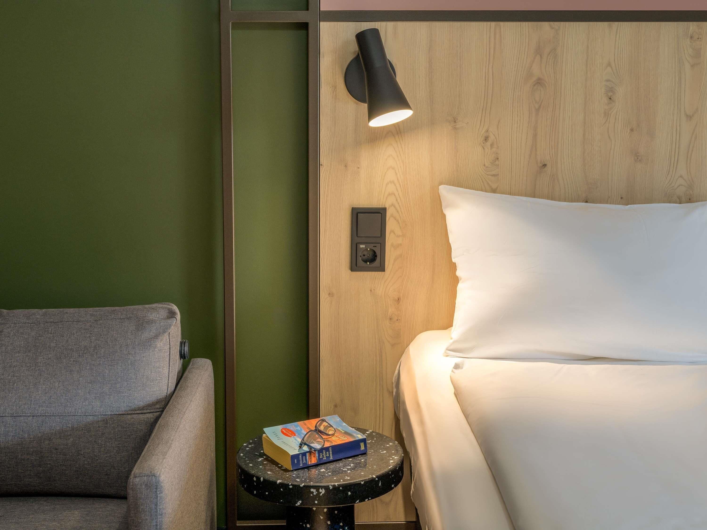 Habitación de hotel con cama, mesita de noche, lámpara de lectura y una pequeña mesa con un libro y gafas.