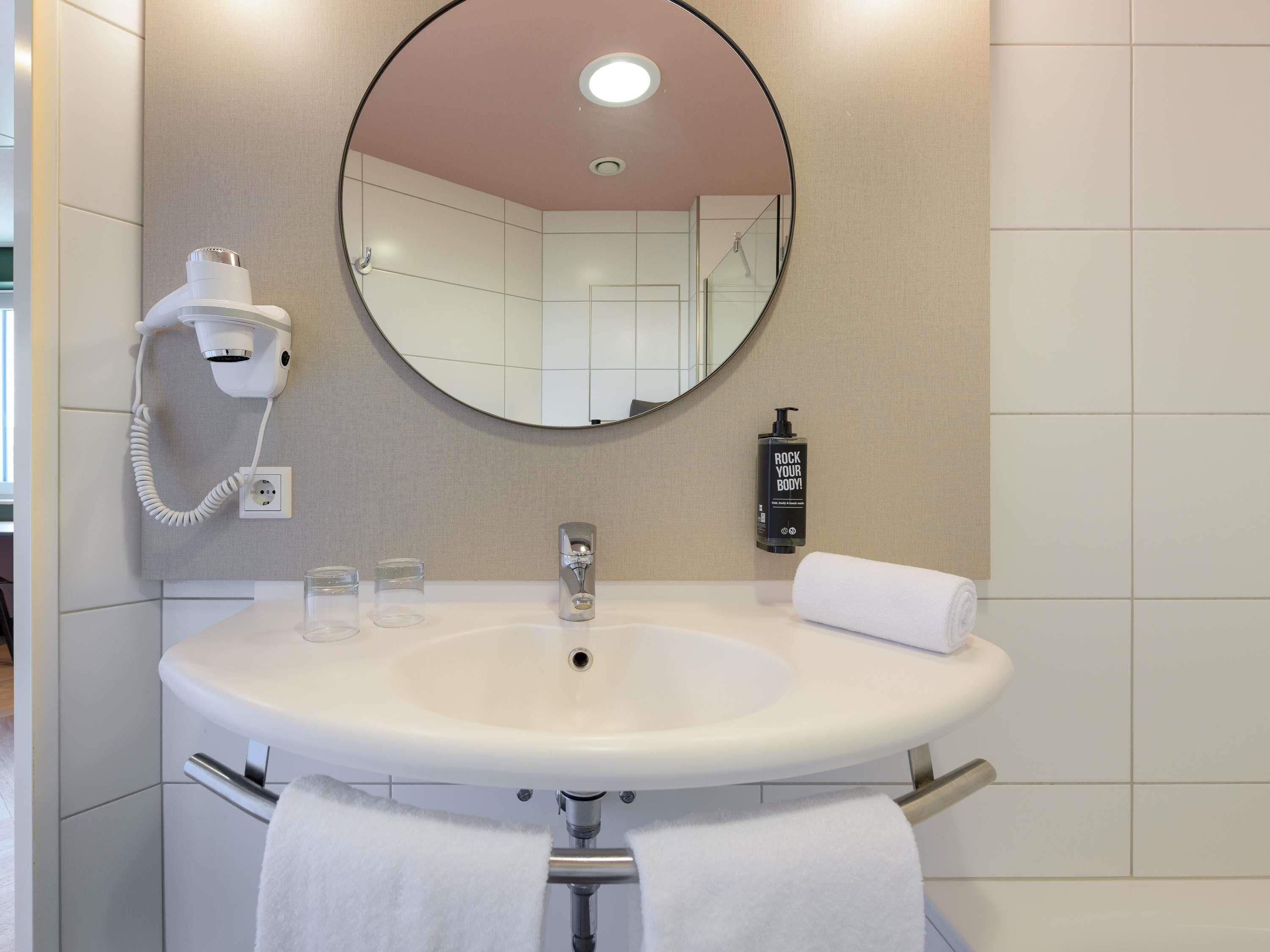 A bathroom with a round mirror, a white sink, a hairdryer, a bottle of body wash, and a towel.