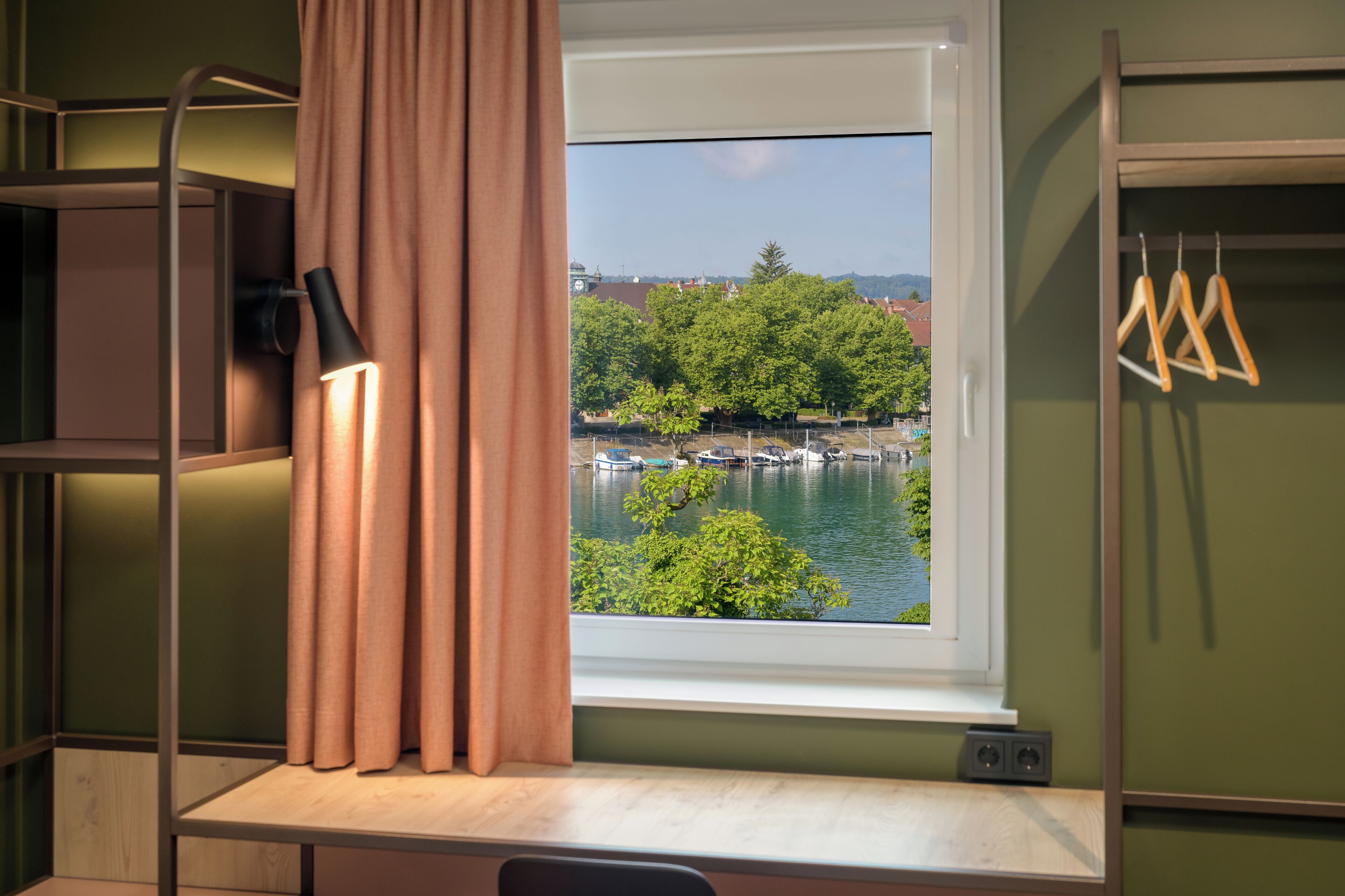 A room with a window overlooking a body of water and greenery, a desk, and a hanging light fixture.