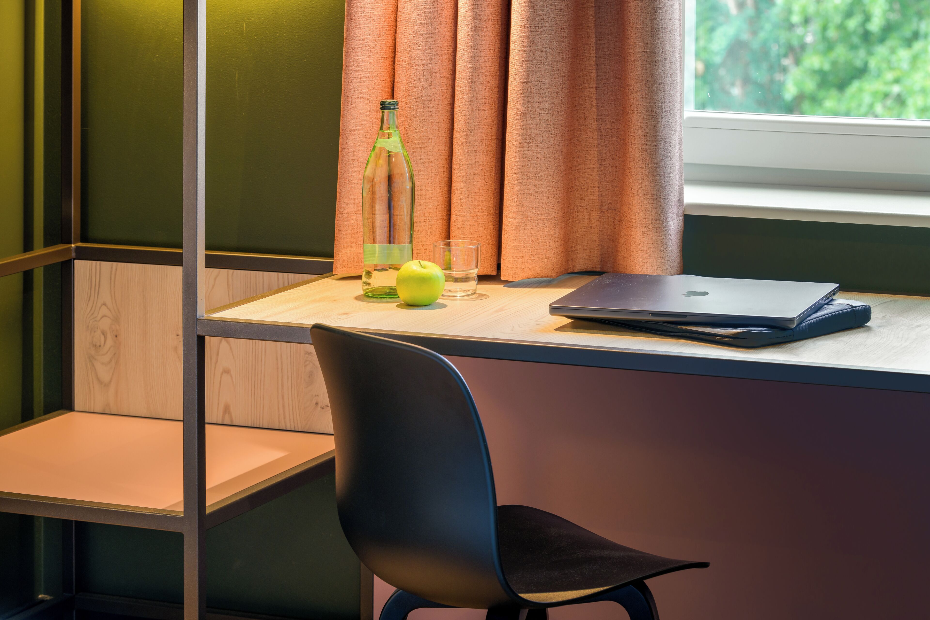 Ein Arbeitsplatz mit Laptop, einer Flasche und einem Apfel auf einem Schreibtisch an einem Fenster.