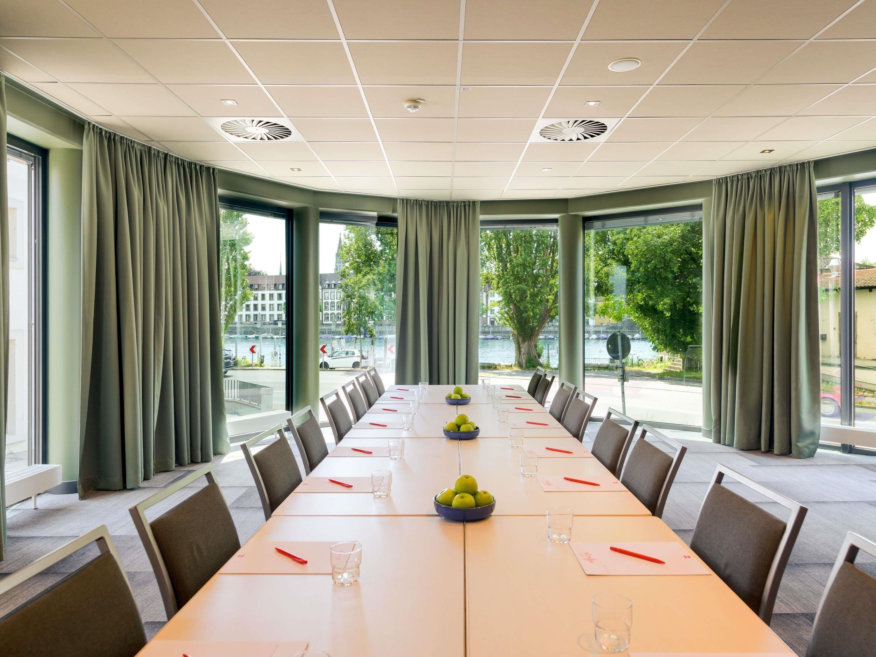 Une salle de conférence avec une longue table, des chaises et de grandes fenêtres équipées de rideaux.