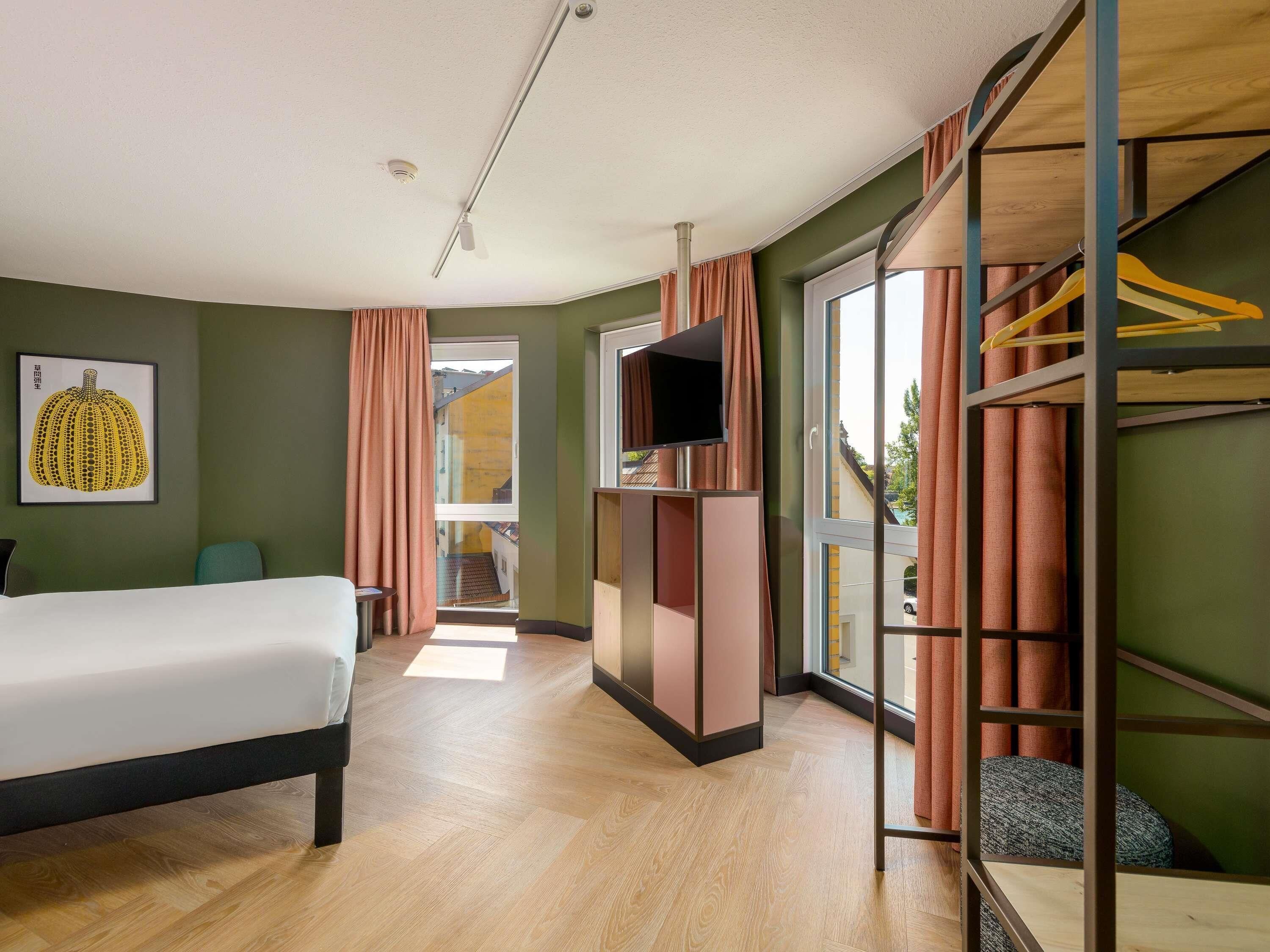 A hotel room with a bed, a TV, a wardrobe, and a view of the outside.