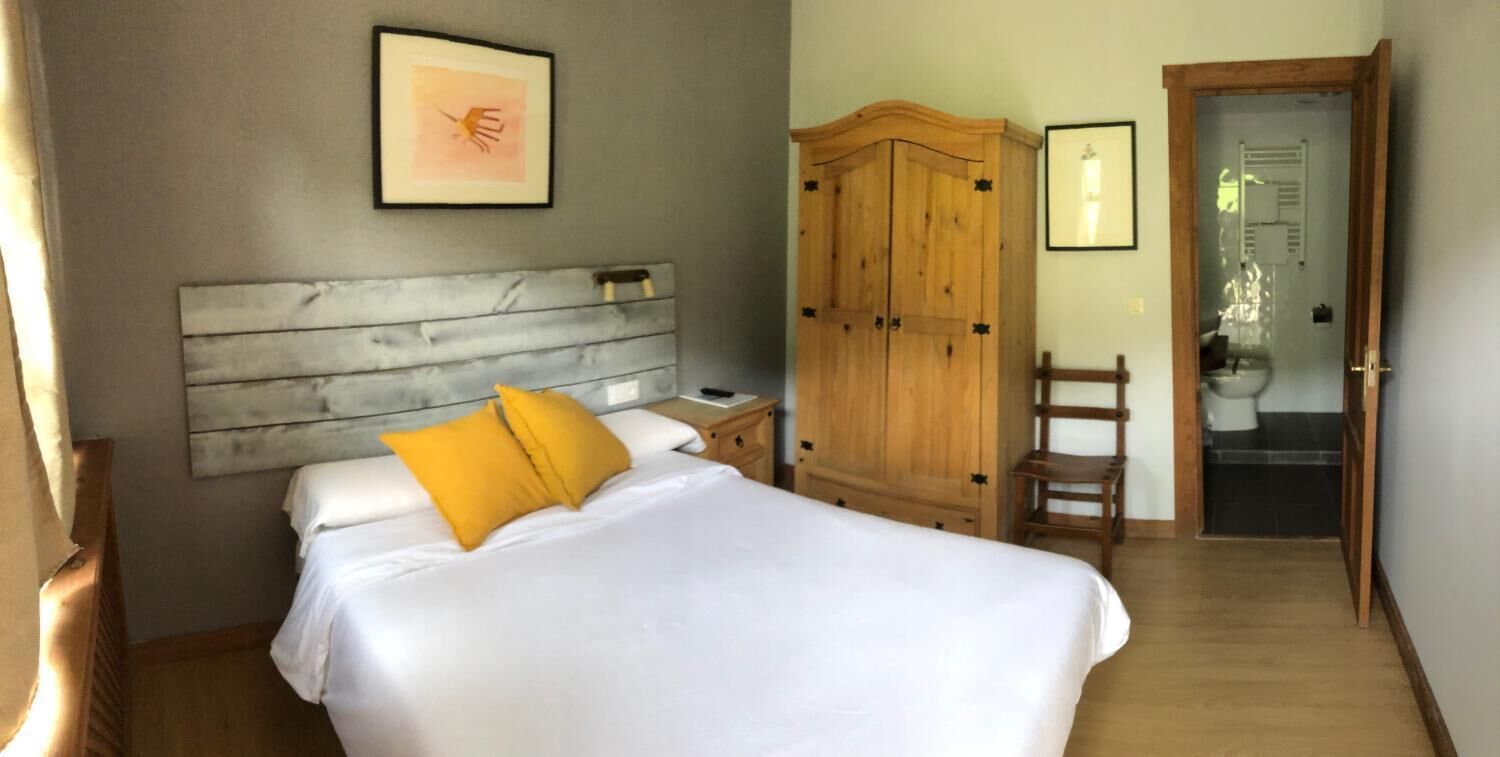 A bedroom with a wooden headboard, a bed with white bedding and yellow pillows, a wooden wardrobe, and a bathroom visible through an open door.